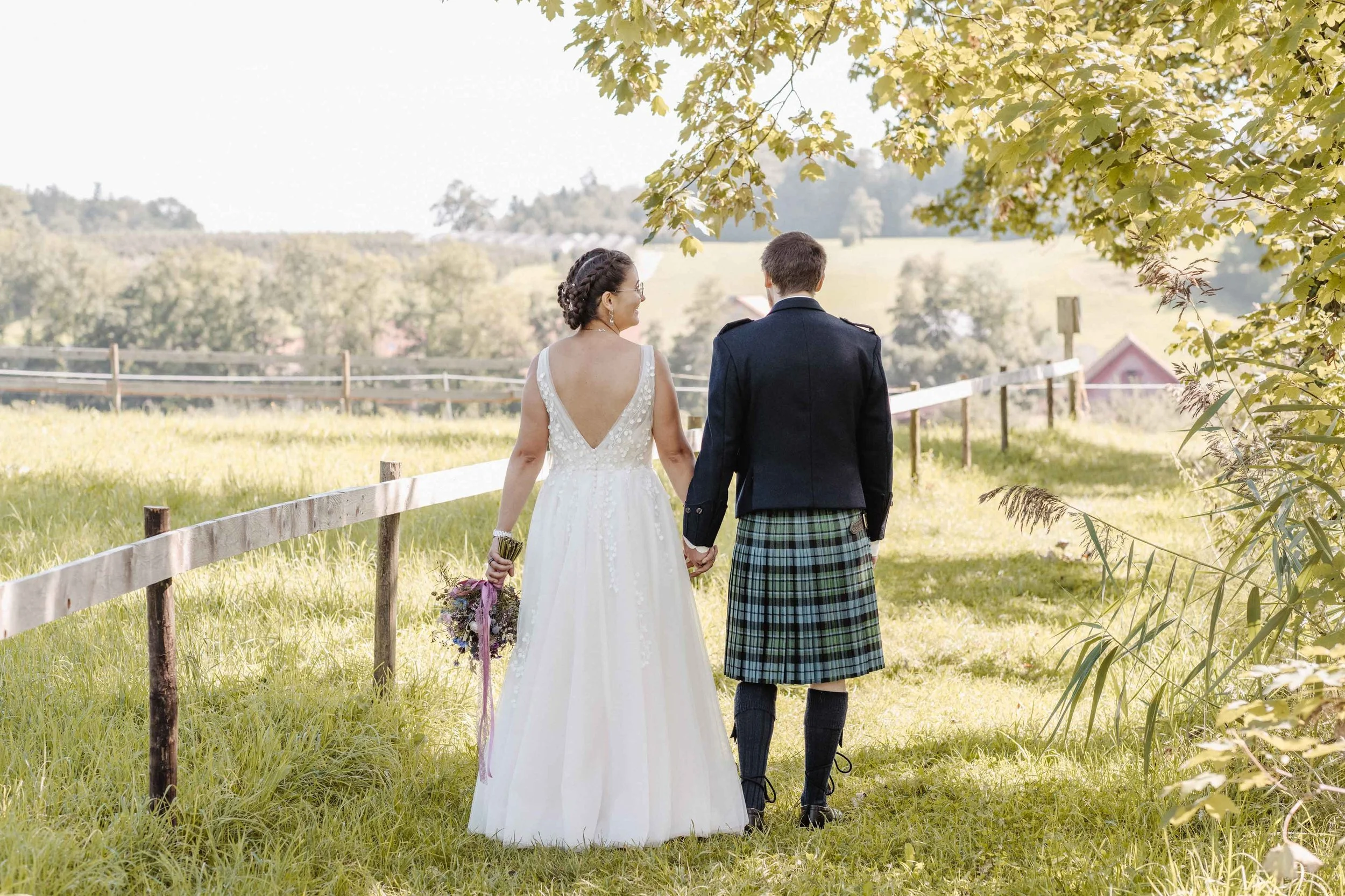 Brautpaar, läuft Hand in Hand auf einer grünen Wiese unter einem Baum, bei Sonnenlicht, im Freien, mit einem Zaun und Bäumen im Hintergrund, während eines sonnigen Tages.
