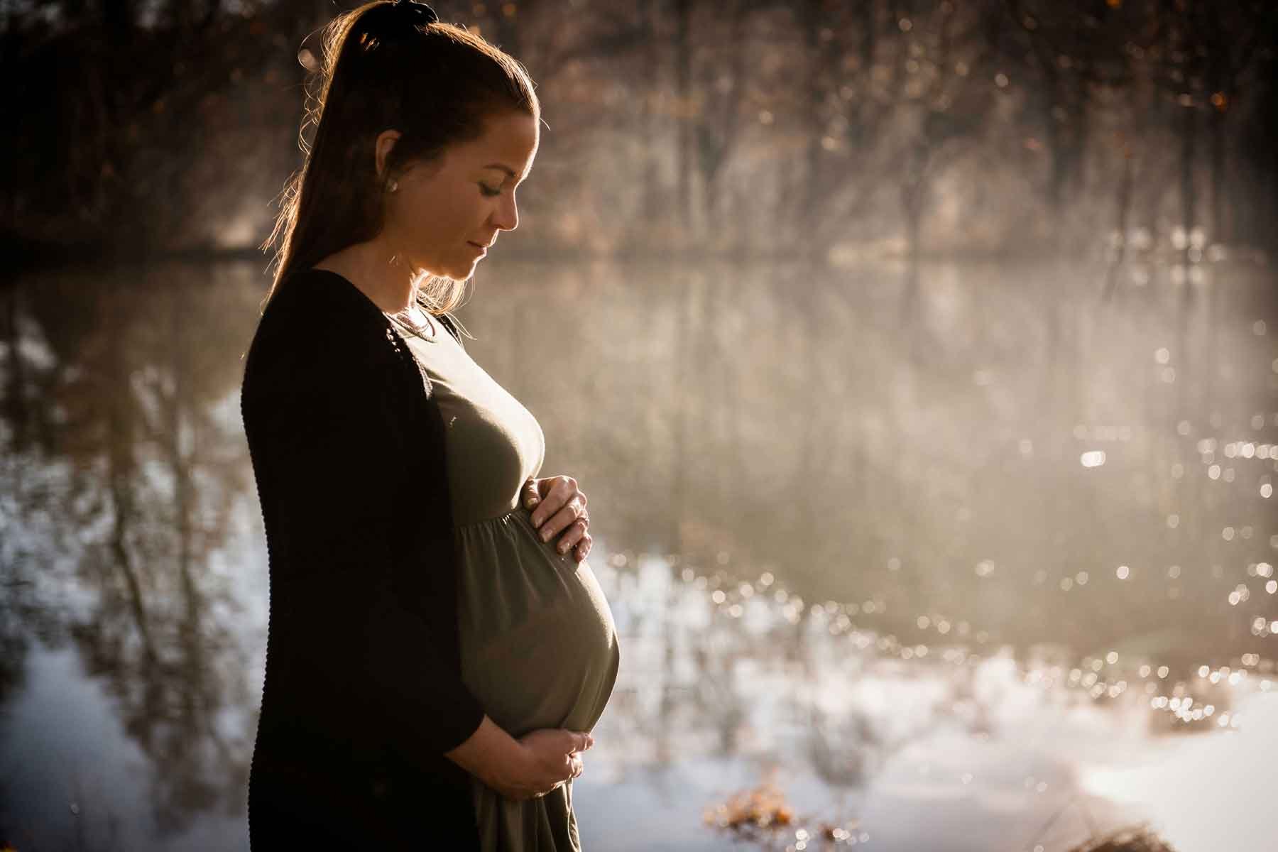 Schwangere Frau im Freien bei Sonnenuntergang an einem Weiher stehend, den Bauch haltend.
