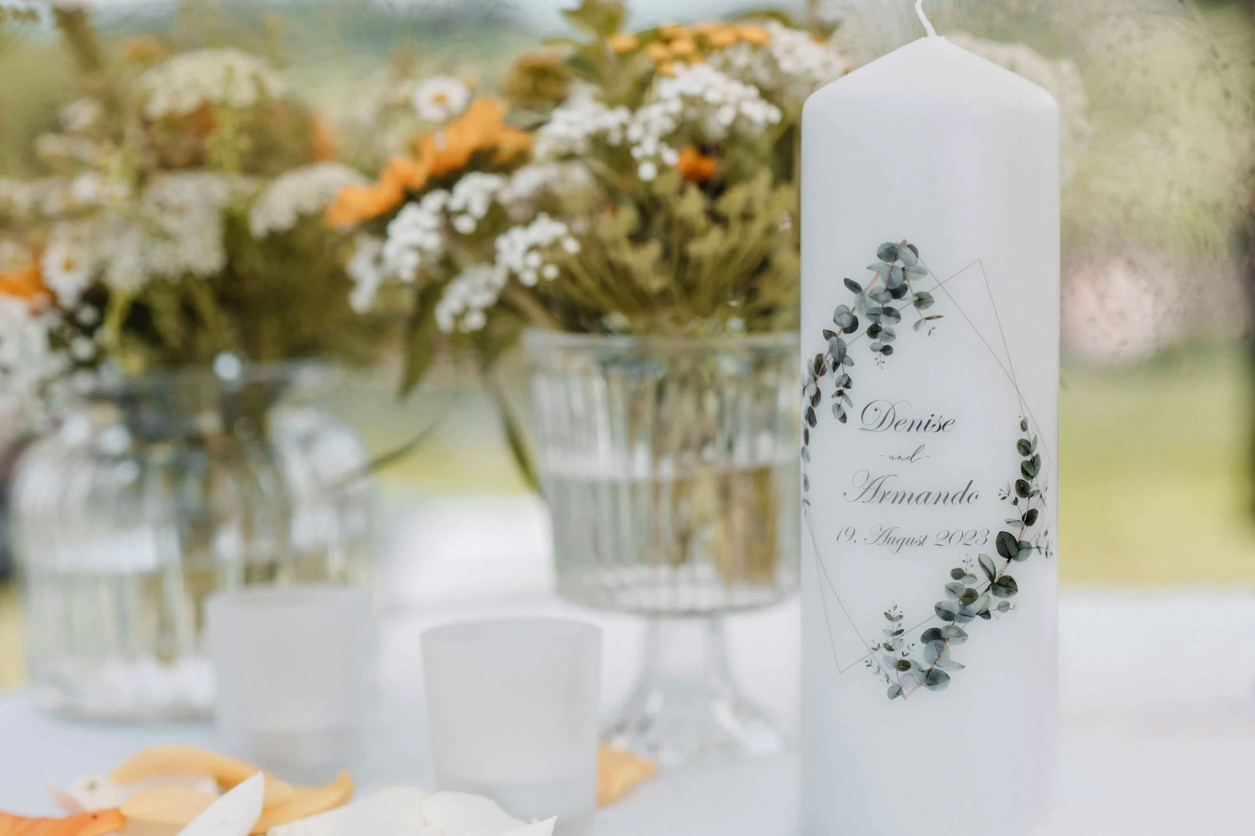 Eine weisse Kerze mit Namen und Datum, umgeben von Eukalyptusblättern, auf einem Tisch bei einer Hochzeit, im Hintergrund Blumen in Vasen.