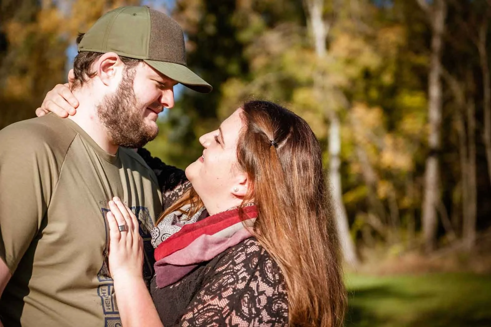 Ein verliebtes Paar steht im Freien, blickt sich tief in die Augen, die Frau hat rote Haare und trägt ein Muster-Shirt mit Schal, der Mann trägt einen Hut und ein braunes T-Shirt, im Hintergrund sind Bäume und Herbstlaub
