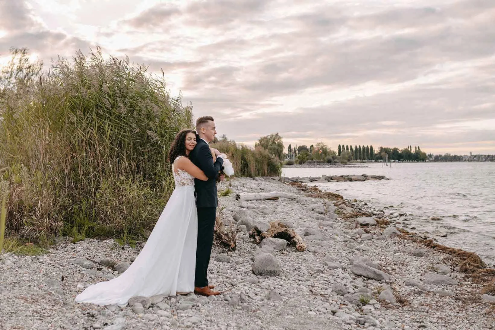 Ein frisch verheiratetes Paar steht am Ufer eines Sees, das Brautpaar umarmt sich. Das Brautkleid ist lang und weiß, der Bräutigam trägt einen dunklen Anzug. Der Himmel ist bewölkt, im Hintergrund sind Bäume und Wasser zu sehen.