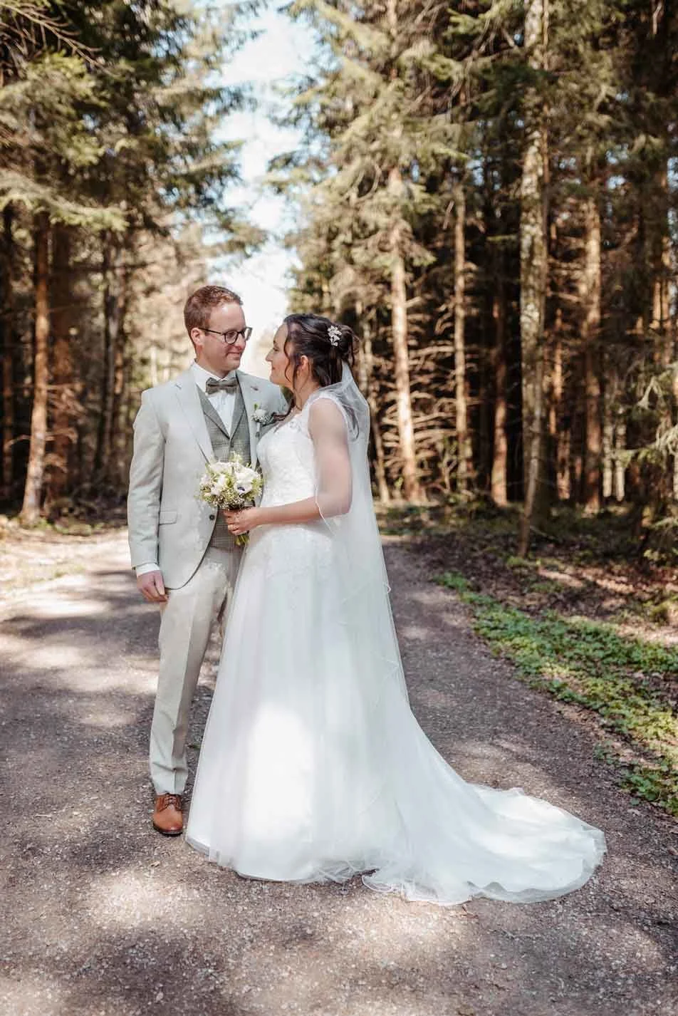 Brautpaar steht in einem Wald, die Braut trägt ein weisses Hochzeitskleid mit Schleier und hält einen Blumenstrauss, der Bräutigam trägt einen hellgrauen Anzug mit Fliege.