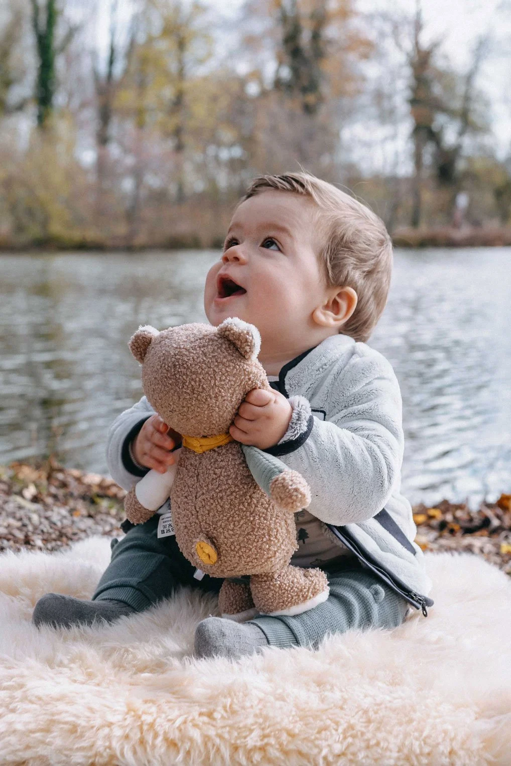 Ein Baby sitzt auf einem flauschigen Teppich am Flussufer im Herbst, hält einen Stoffbär und schaut nach oben.