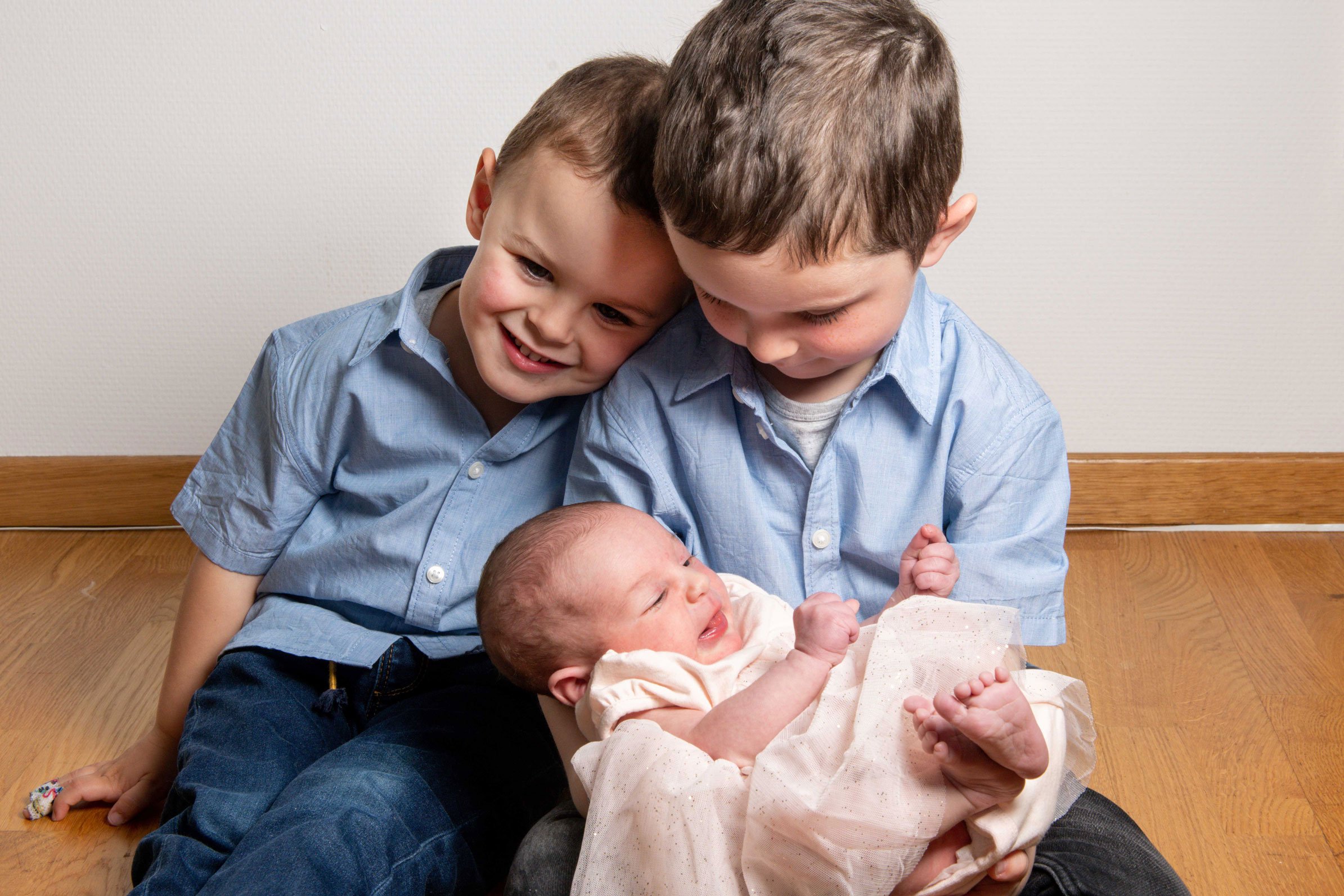 Zwei Kinder sitzen auf dem Boden und halten ein Baby in den Armen. Alle tragen blaue Hemden, das Baby ist in ein helles Kleid gehüllt.