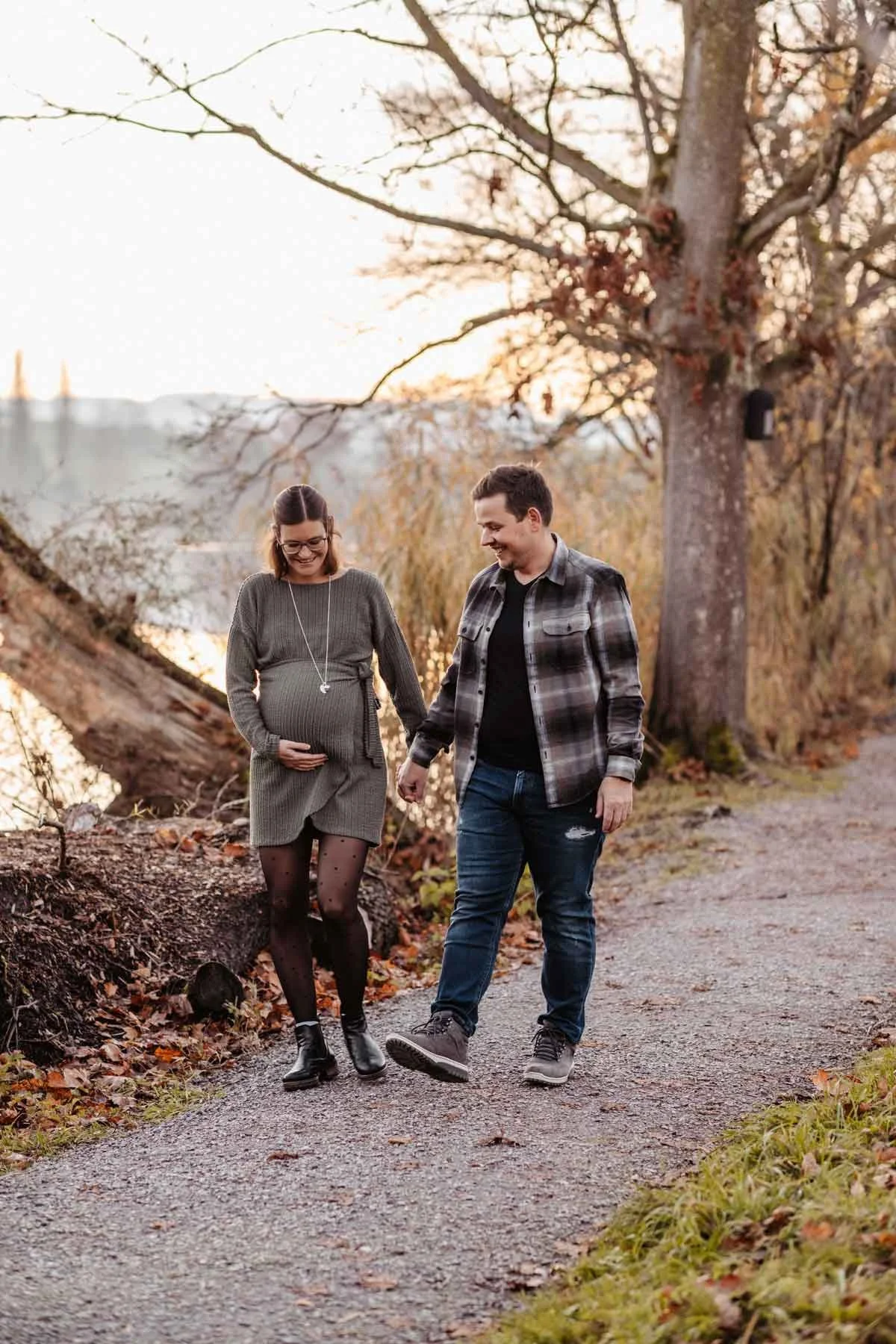 Ein schwangere Frau und ein Mann gehen Hand in Hand auf einem Weg entlang eines Weihers im herbstlichen Park, die Frau hält ihren Bauch. Es ist Sonnenuntergang, und die Bäume sind herbstlich gefärbt.