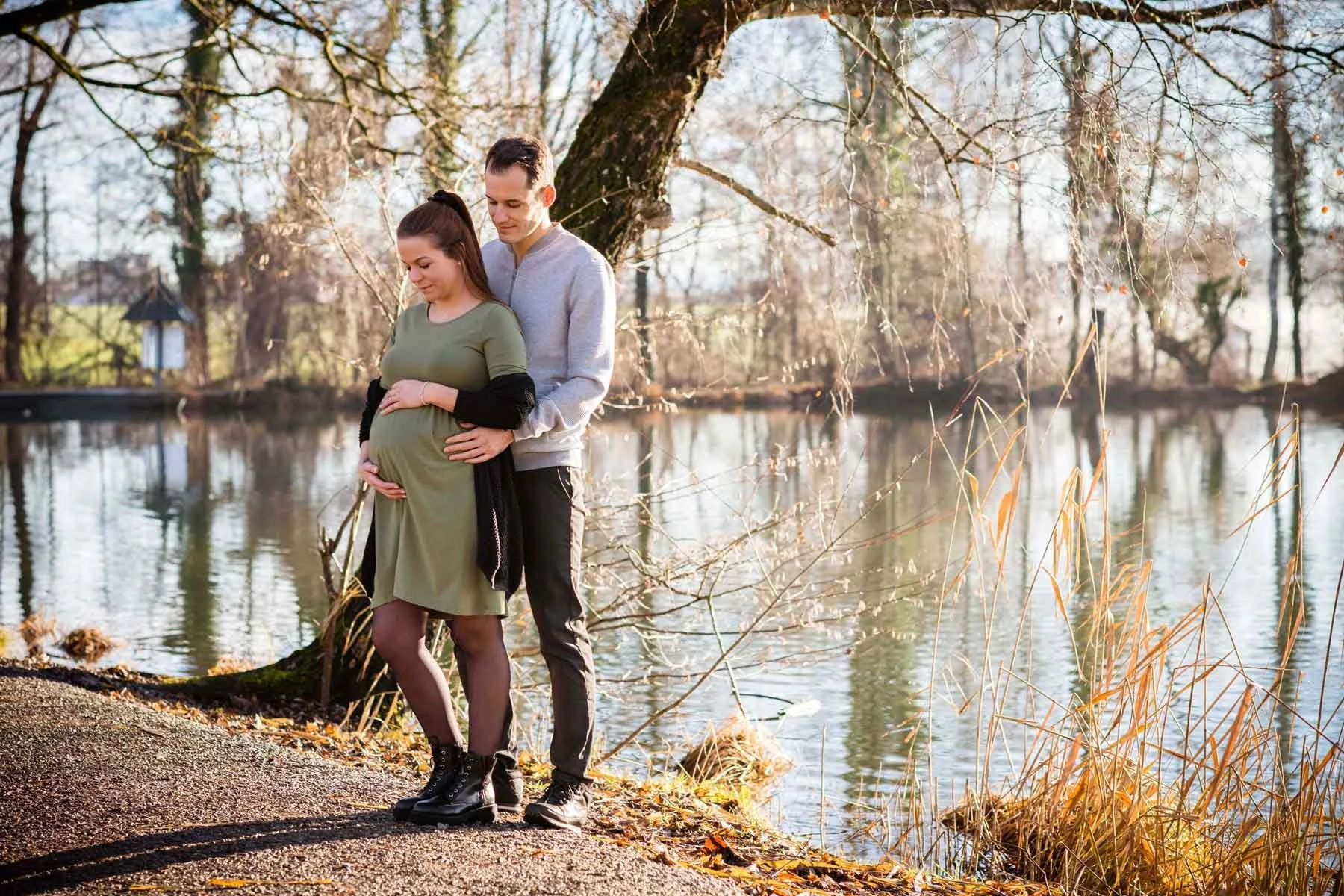 Ein schwangeres Paar steht am Ufer eines Sees im Herbst, umgeben von Bäumen ohne Blätter, bei Sonnenlicht, outdoor, romantisch.