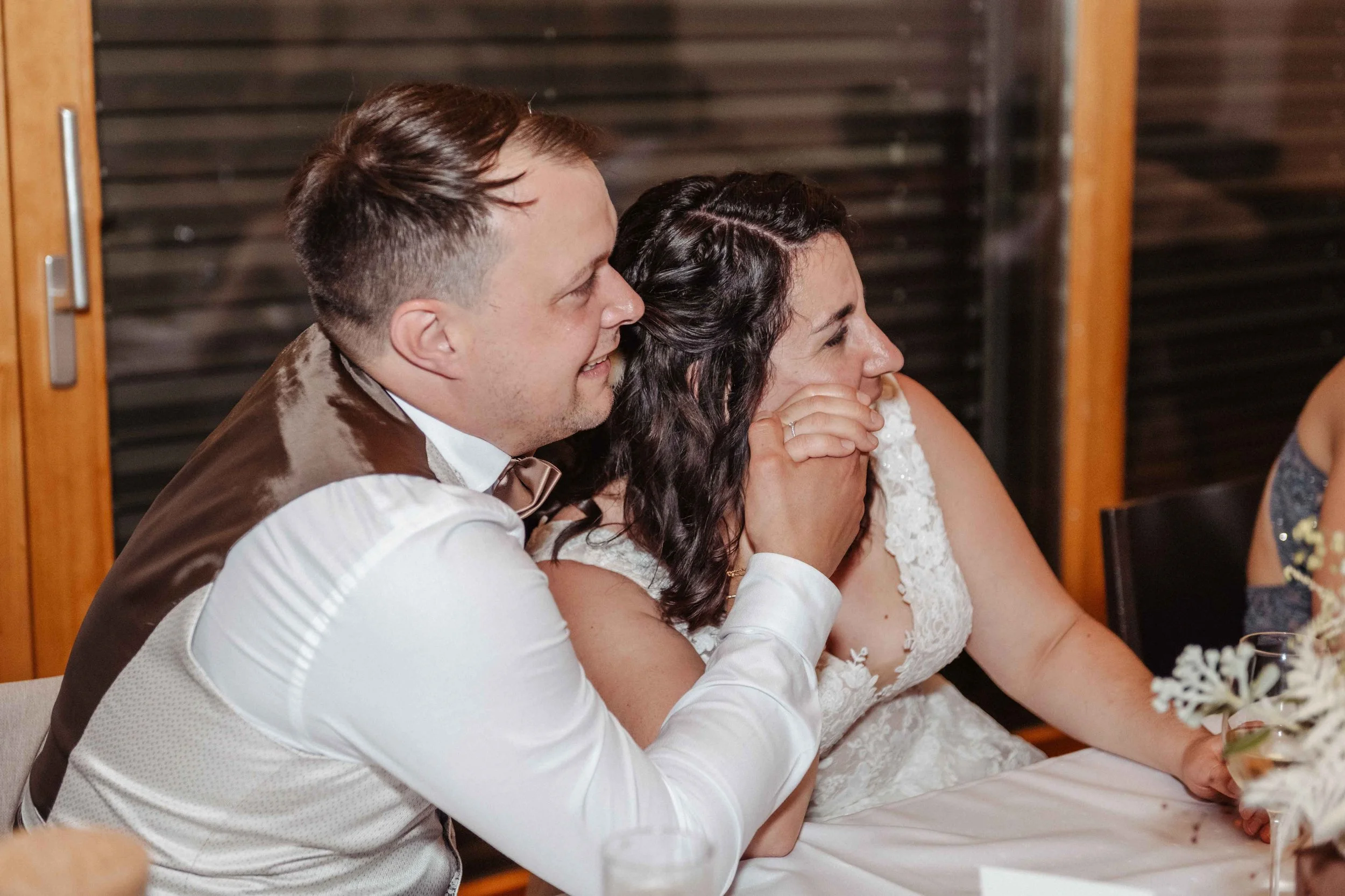 Brautpaar sitzt glücklich und lachend am Tisch, die Braut trägt ein weißes Hochzeitskleid, der Bräutigam trägt ein Hemd mit Weste.