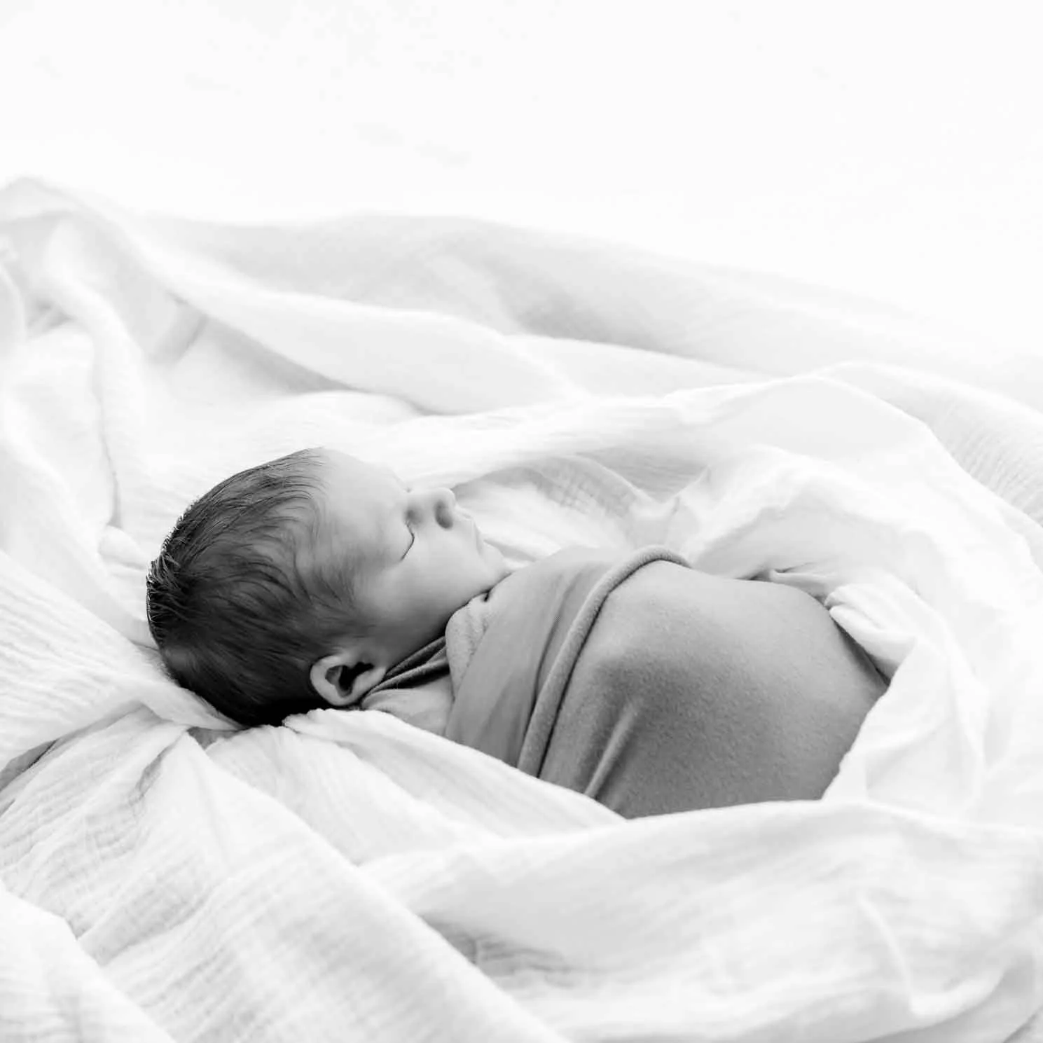 Schläfriger Neugeborenes liegt auf einem Bett und ist in eine Decke gehüllt, schwarz-weisse Aufnahme.