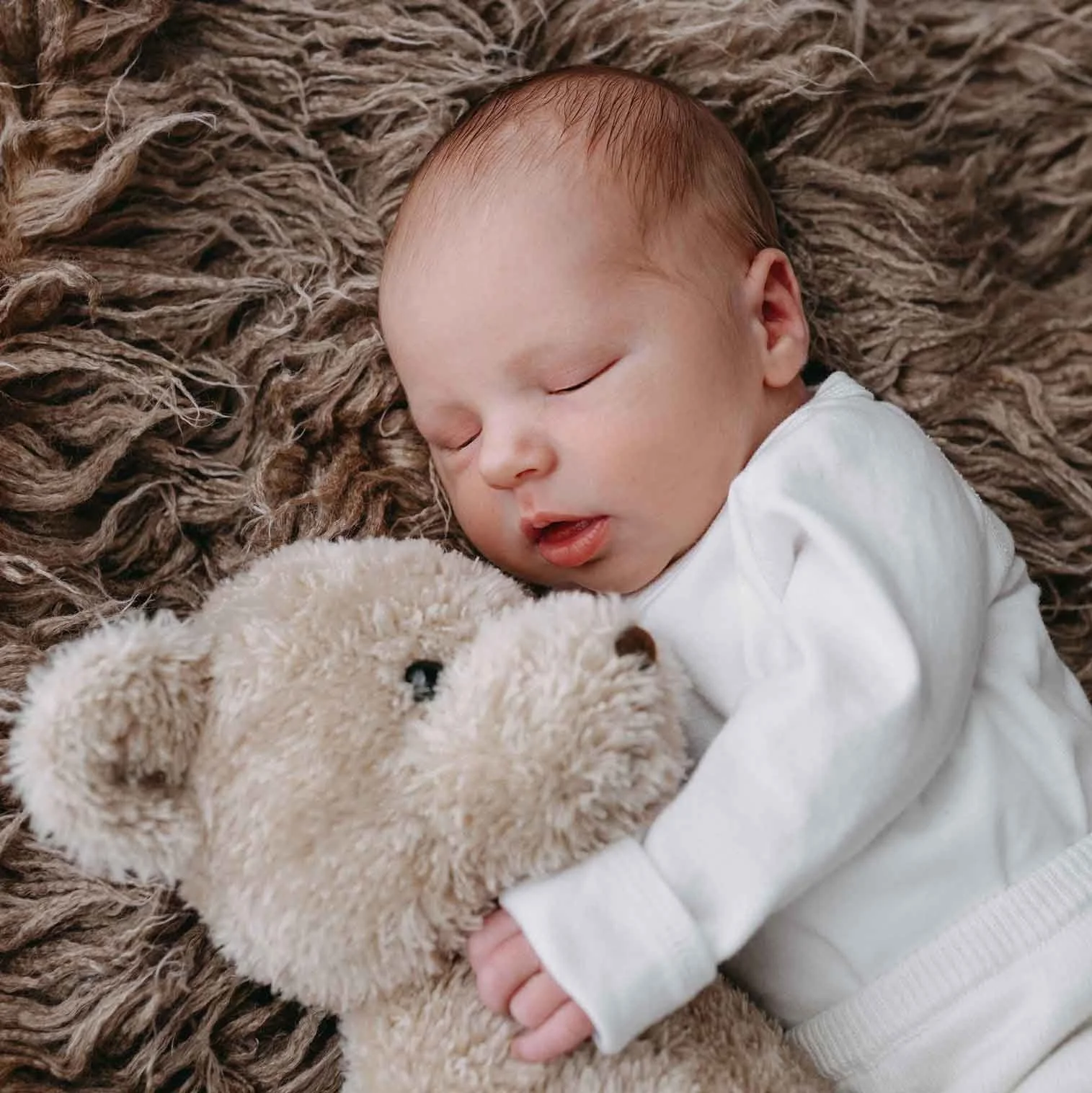 Kleines Baby schläft auf einer weichen, braunen Decke und hält einen beigen Plüschbären