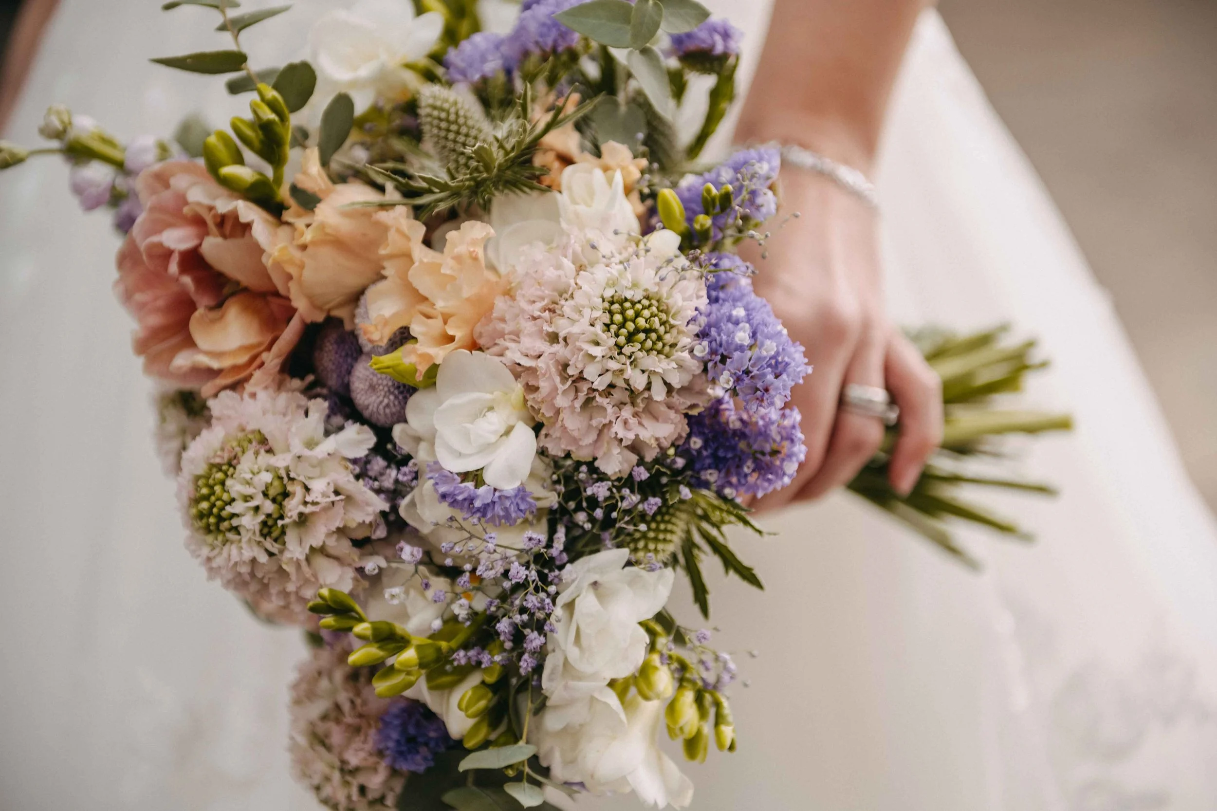 Braut hält Brautstrauss aus verschiedenen Blumen in Pastellfarben, darunter Pfirsich, Weiß und Lila.