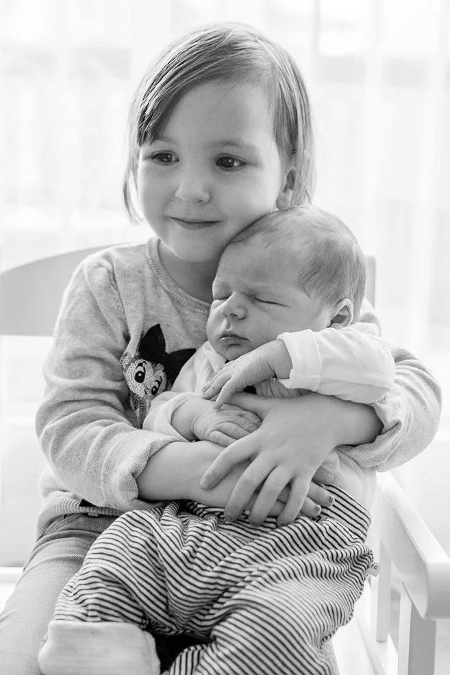 Ein Mädchen hält das Baby im Arm, beide sitzen auf einem Stuhl vor einem Fenster, in Schwarz-Weiss aufgenommen.