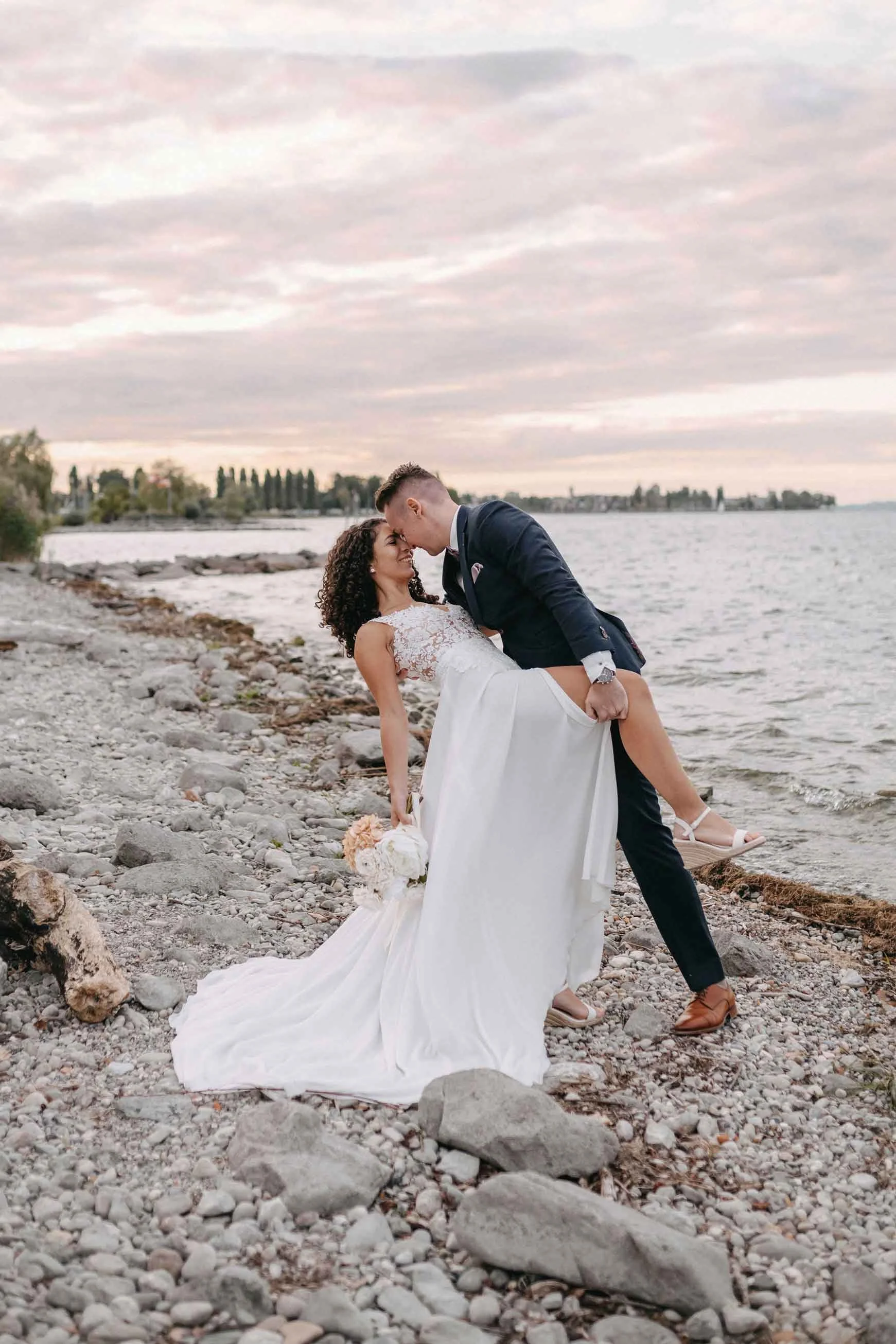 Hochzeitspaar am Strand, der Mann hebt die Braut auf und küsst sie, im Hintergrund Wasser und Himmel bei Sonnenuntergang.
