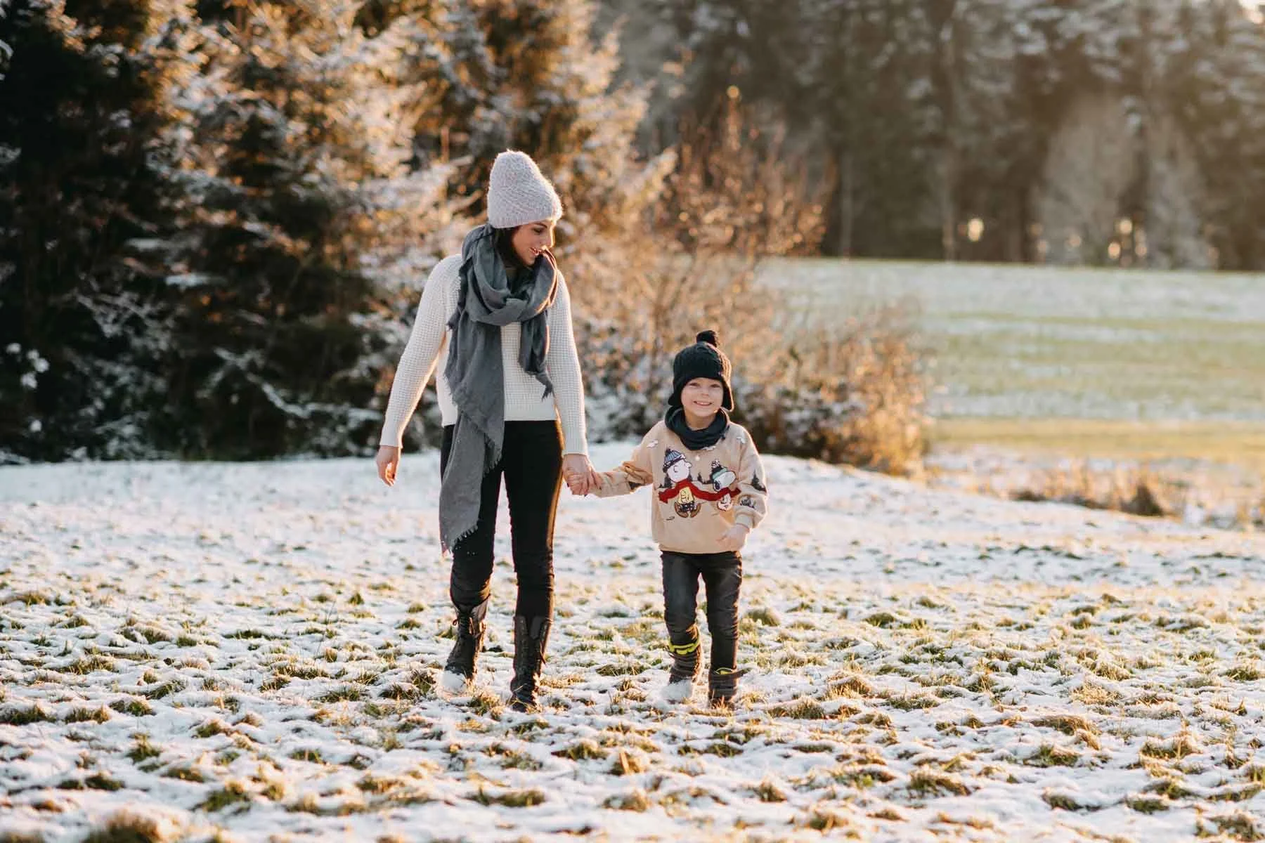 Mama mit Kind gehen im Schnee spazieren, umgeben von Bäumen und Wiesen.