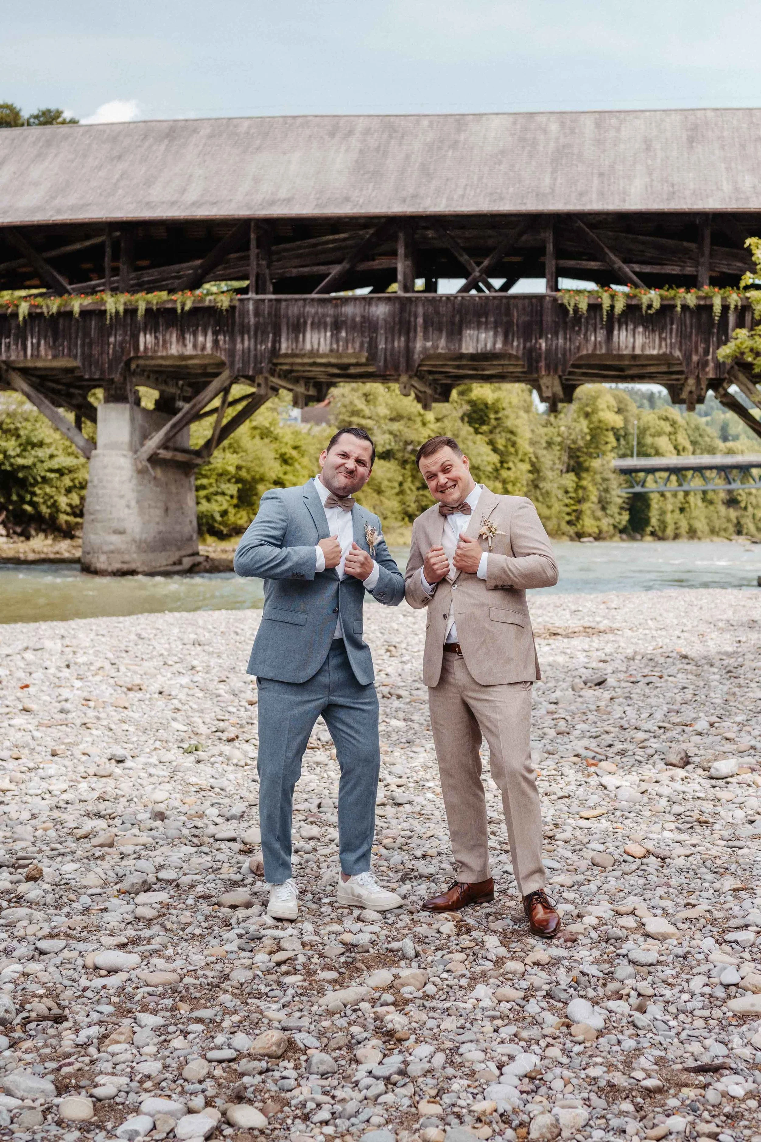 Zwei Männer in Anzügen posieren lachend am Flussufer vor einer historischen Holzbrücke.