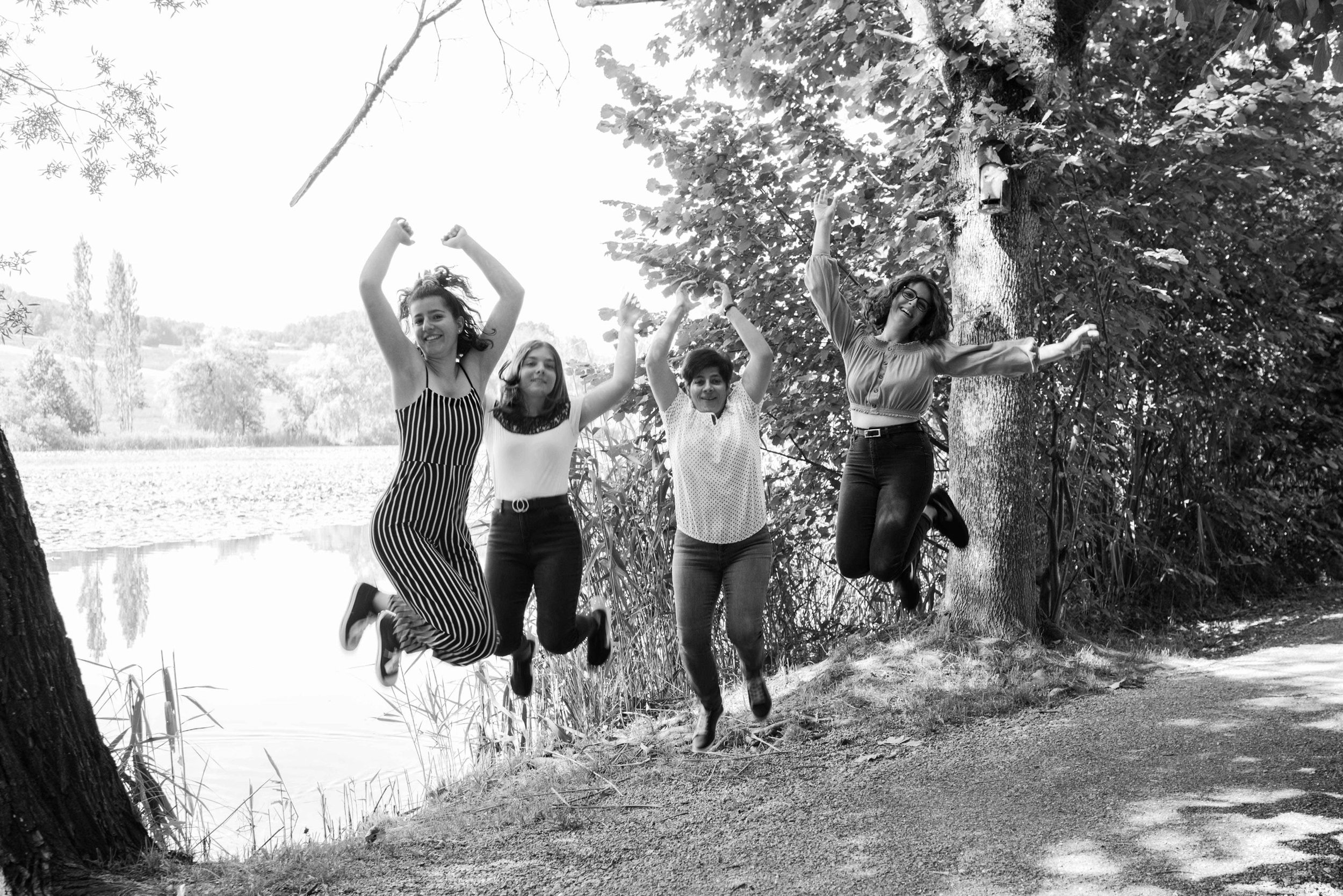Vier Frauen springen fröhlich in der Luft auf einem Waldweg neben einem See.