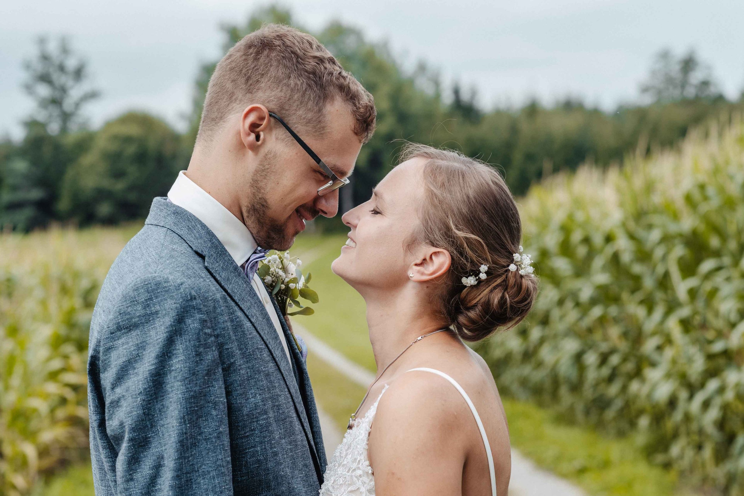 Brautpaar steht sich im Freien sehr nahe gegenüber, lachend und verliebt, umgeben von einer grünen Landschaft.