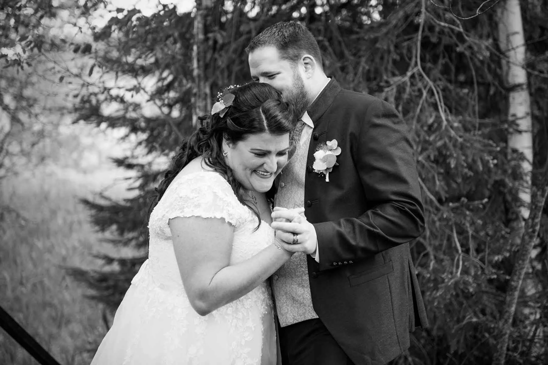 Braut und Bräutigam tanzen bei einer Hochzeit im Freien, lachen und halten Händchen, im Hintergrund Bäume.