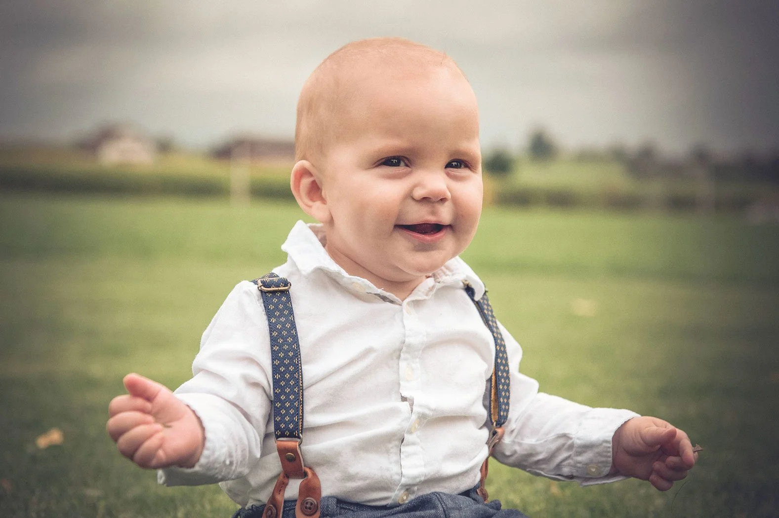 Lächelnder Babyjunge im weissen Hemd mit Hosenträgern sitzt auf einer Wiese im Freien.