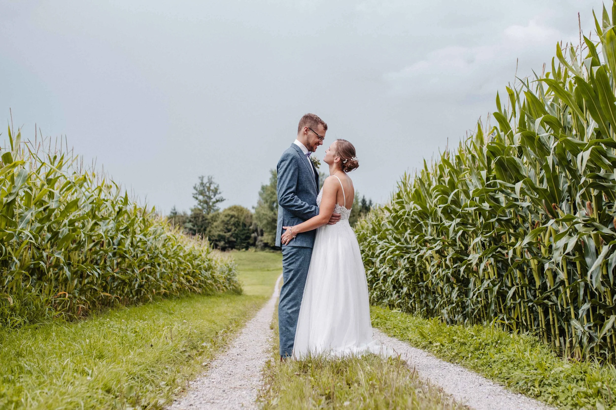 Ein frisch verheiratetes Paar auf einer landwirtschaftlichen Strasse, umgeben von Maisfeldern, bei bewölktem Himmel.