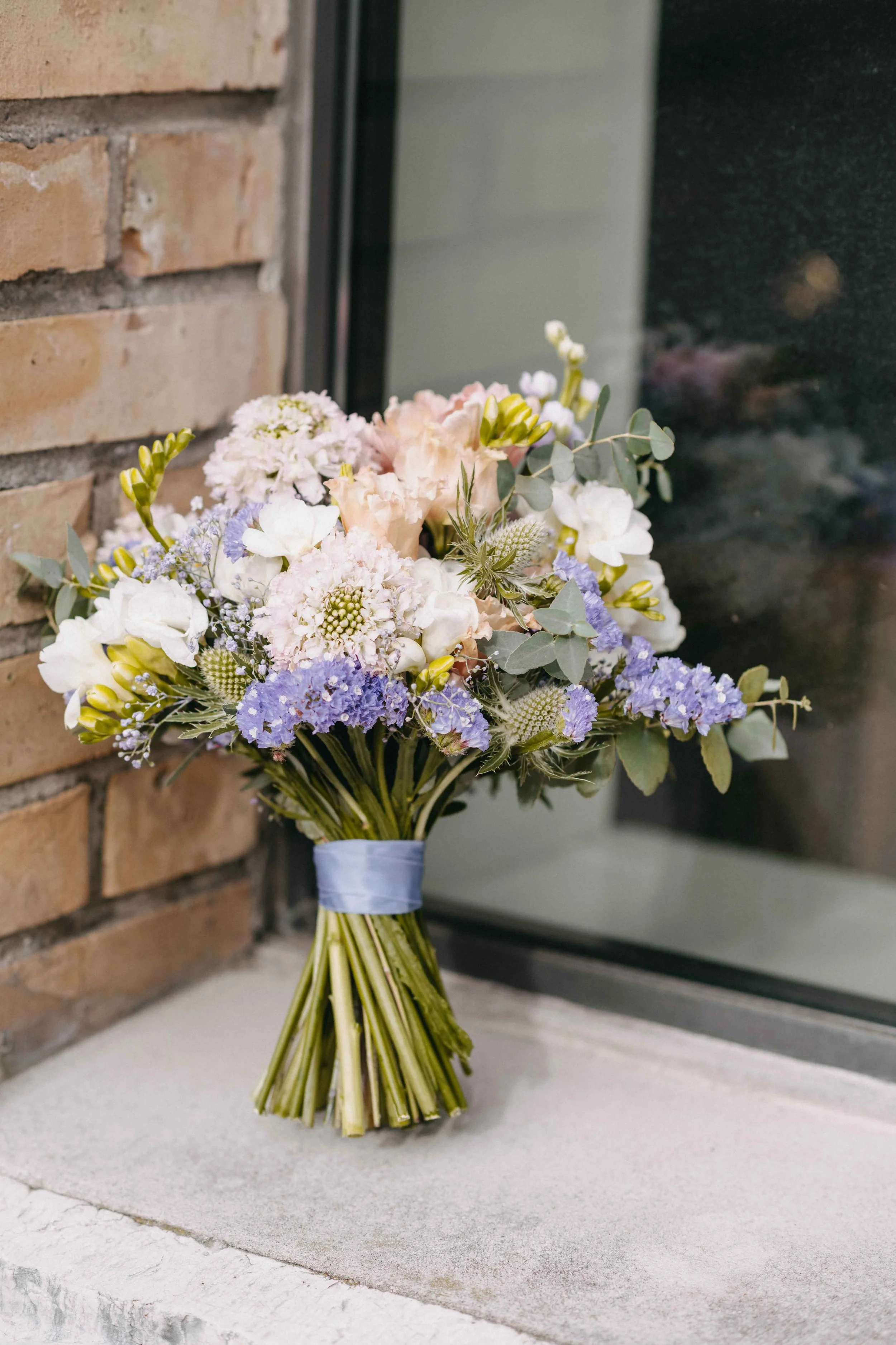 Brautstrauss aus weissen, lila und blass-pinken Blumen steht auf einer Fensterbank neben einer Backsteinwand.