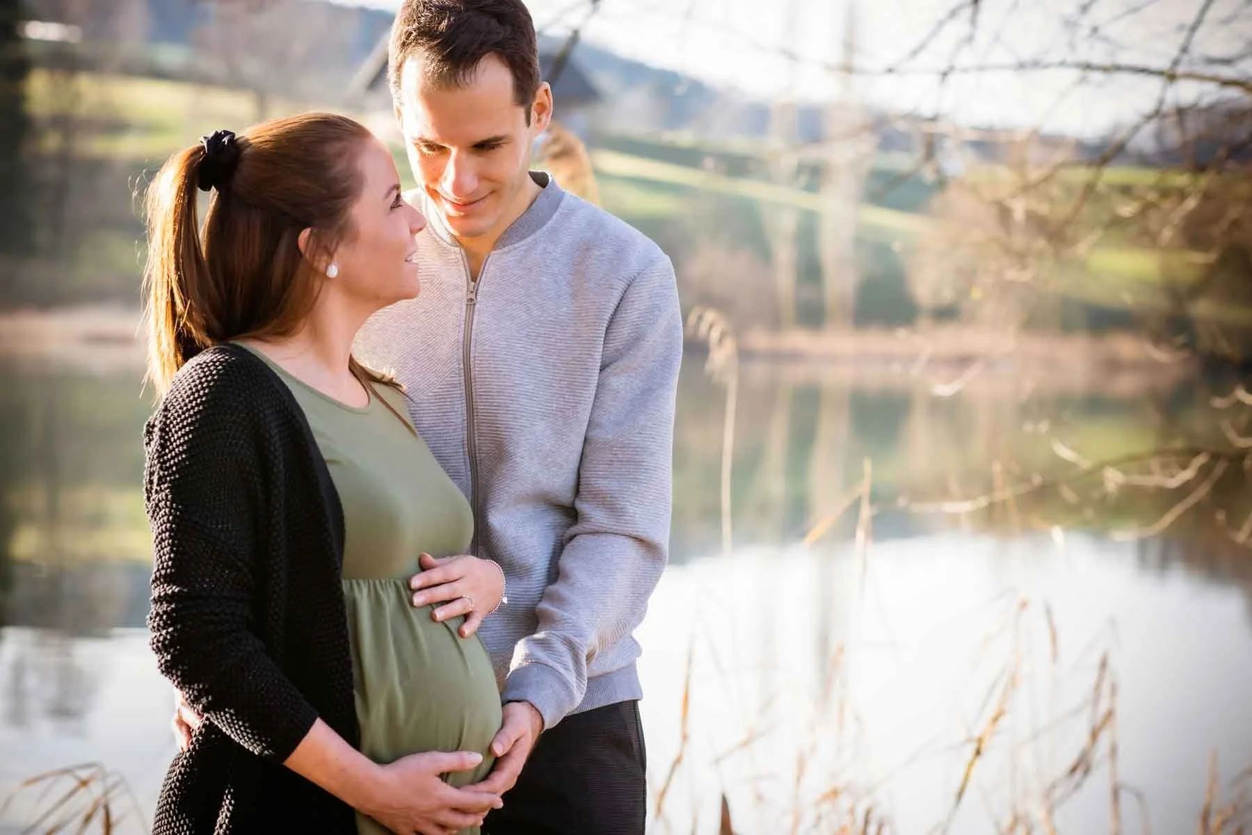 Ein schwangeres Paar steht am Fluss und blickt sich liebevoll an.