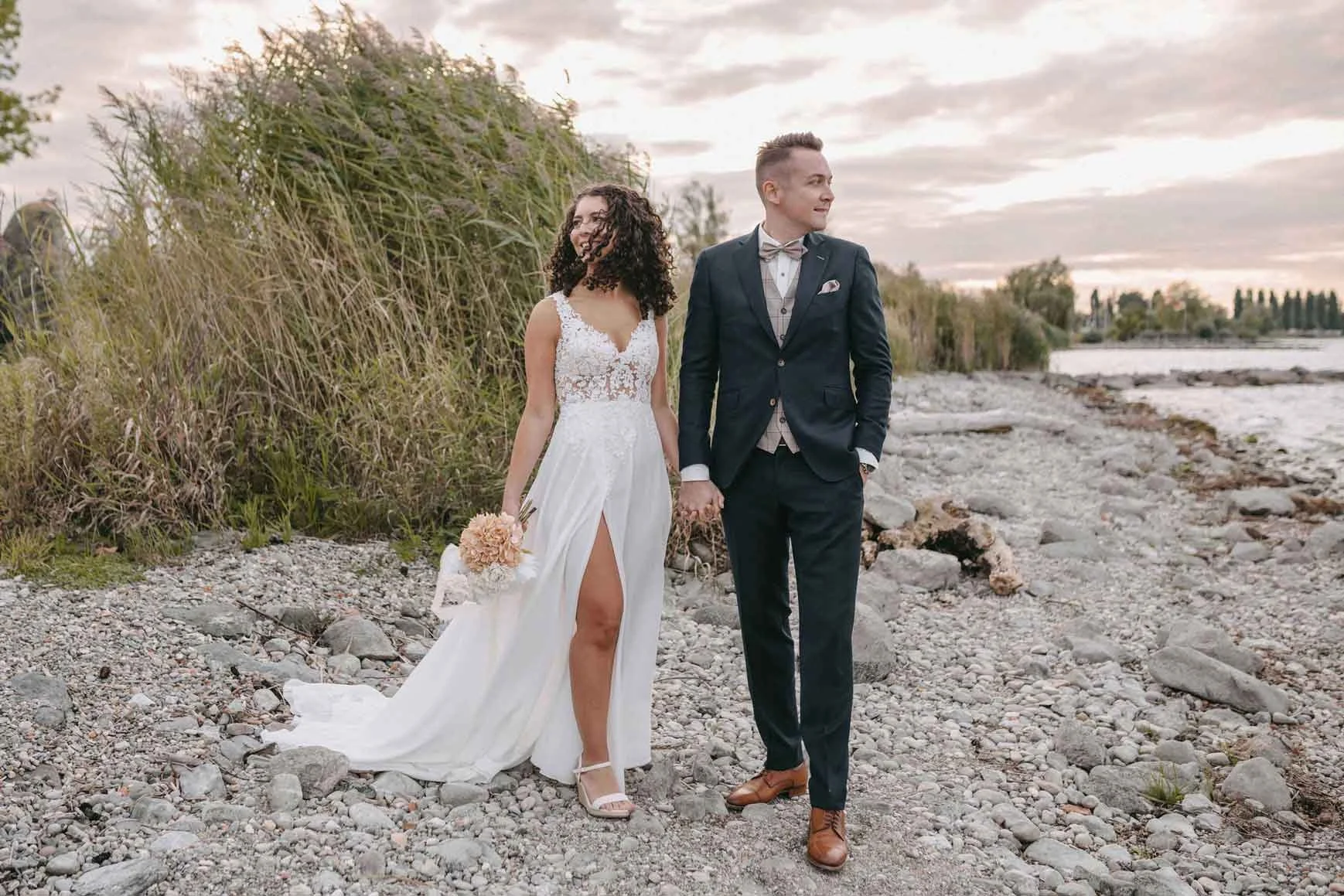 Brautpaar in Hochzeitskleidung spaziert am Strand entlang, hält Händchen, bei Sonnenuntergang, im Freien, romantisch.