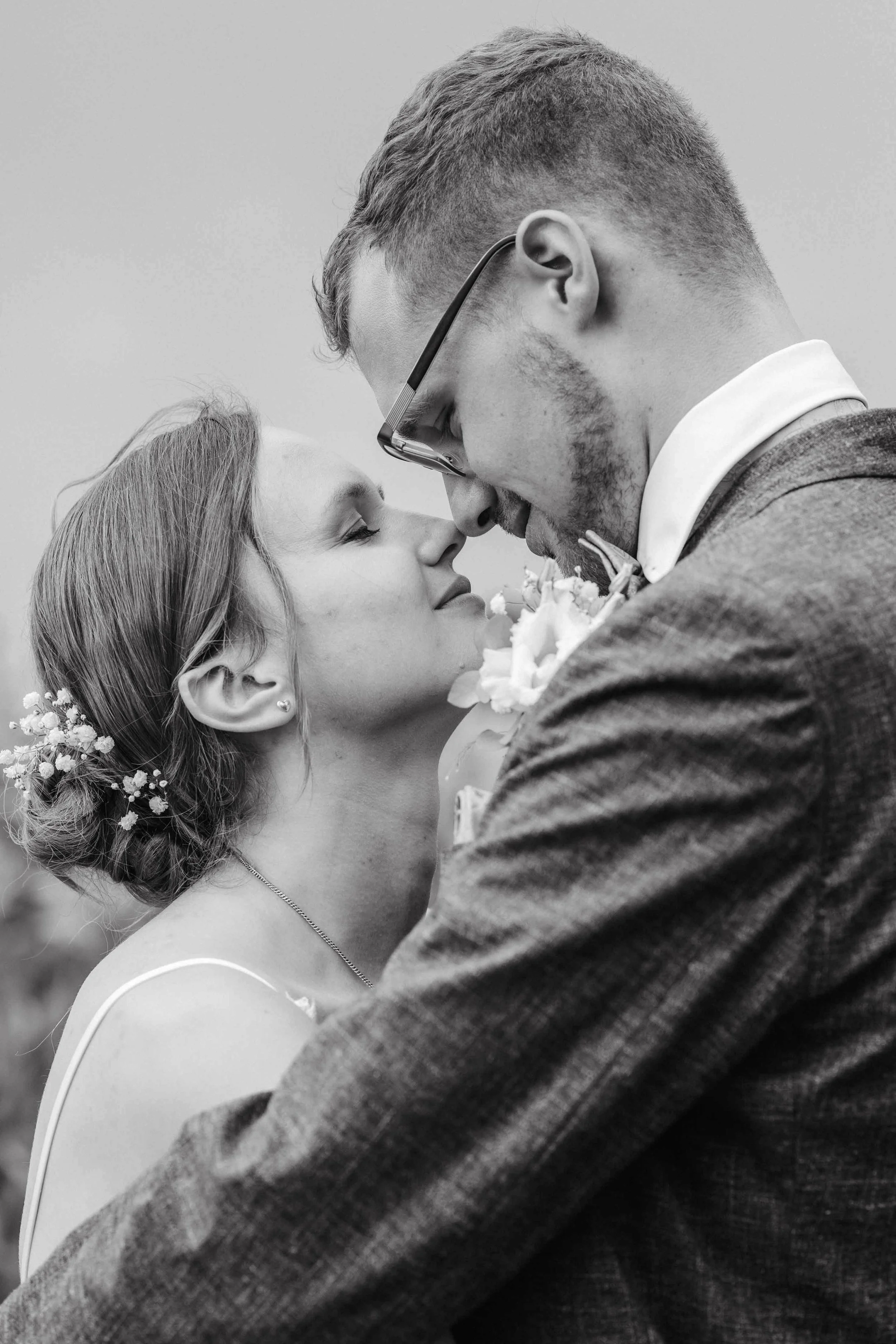 Brautpaar beim Paarshooting, die Frau trägt ein Hochzeitskleid mit Blumen im Haar, der Mann trägt einen Anzug, sie stehen sich nah und berühren die Stirnen, schwarz-weiß Foto.