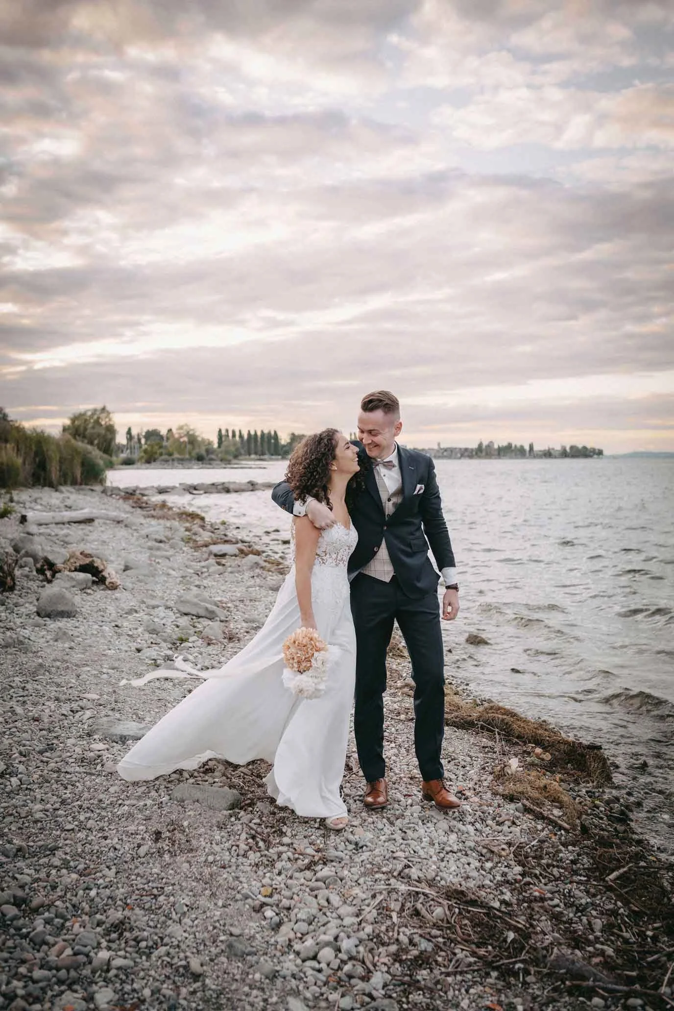Brautpaar am Ufer eines Sees bei Sonnenuntergang, lachend und sich umarmend.