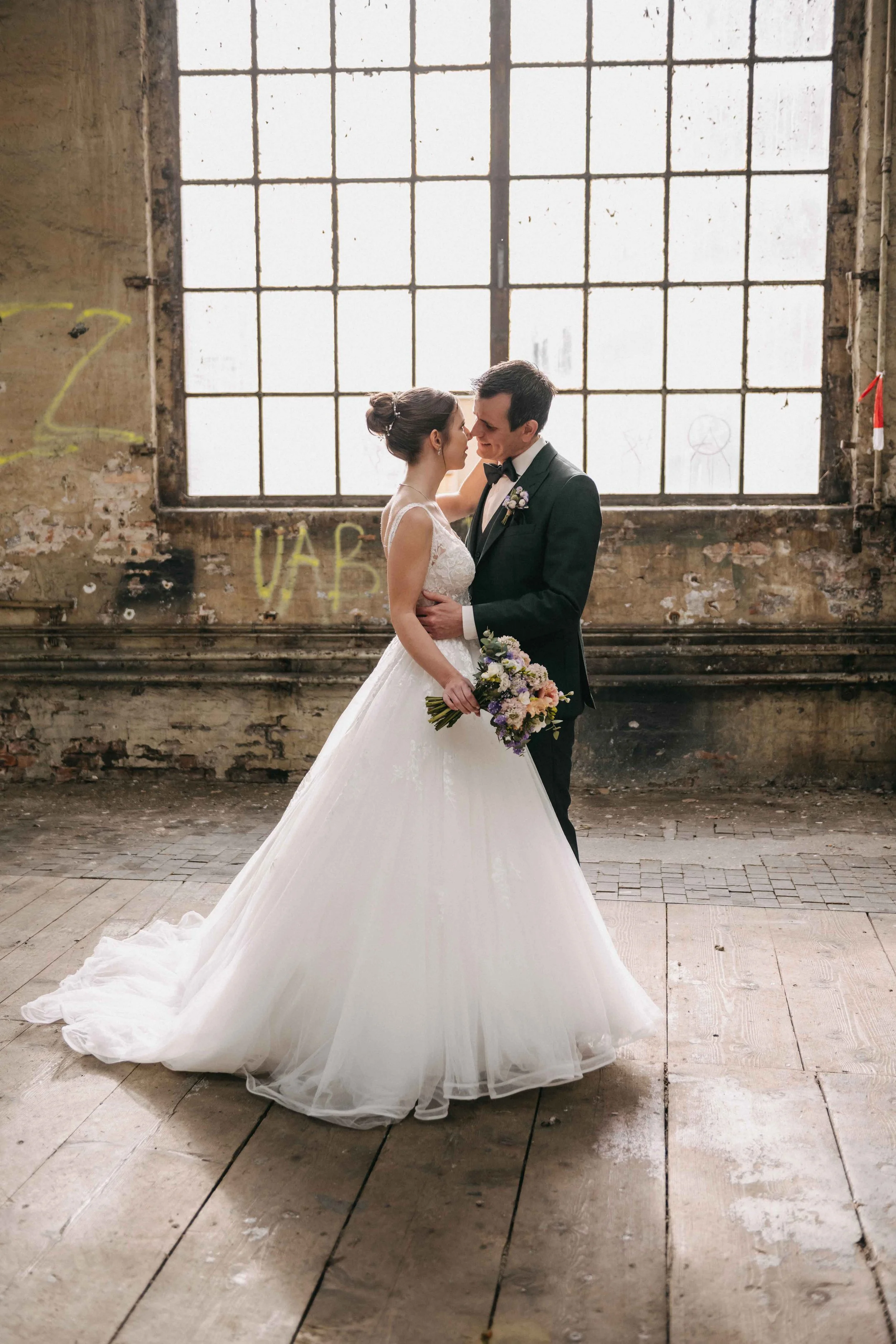 Brautpaar steht in einem rustikalen, alten industriellem Raum, das Paar ist im Arm und blickt sich liebevoll an. Das Brautkleid ist lang und weiss, die Braut hält einen Blumenstrauss. Die Szene ist romantic und intimitiv.