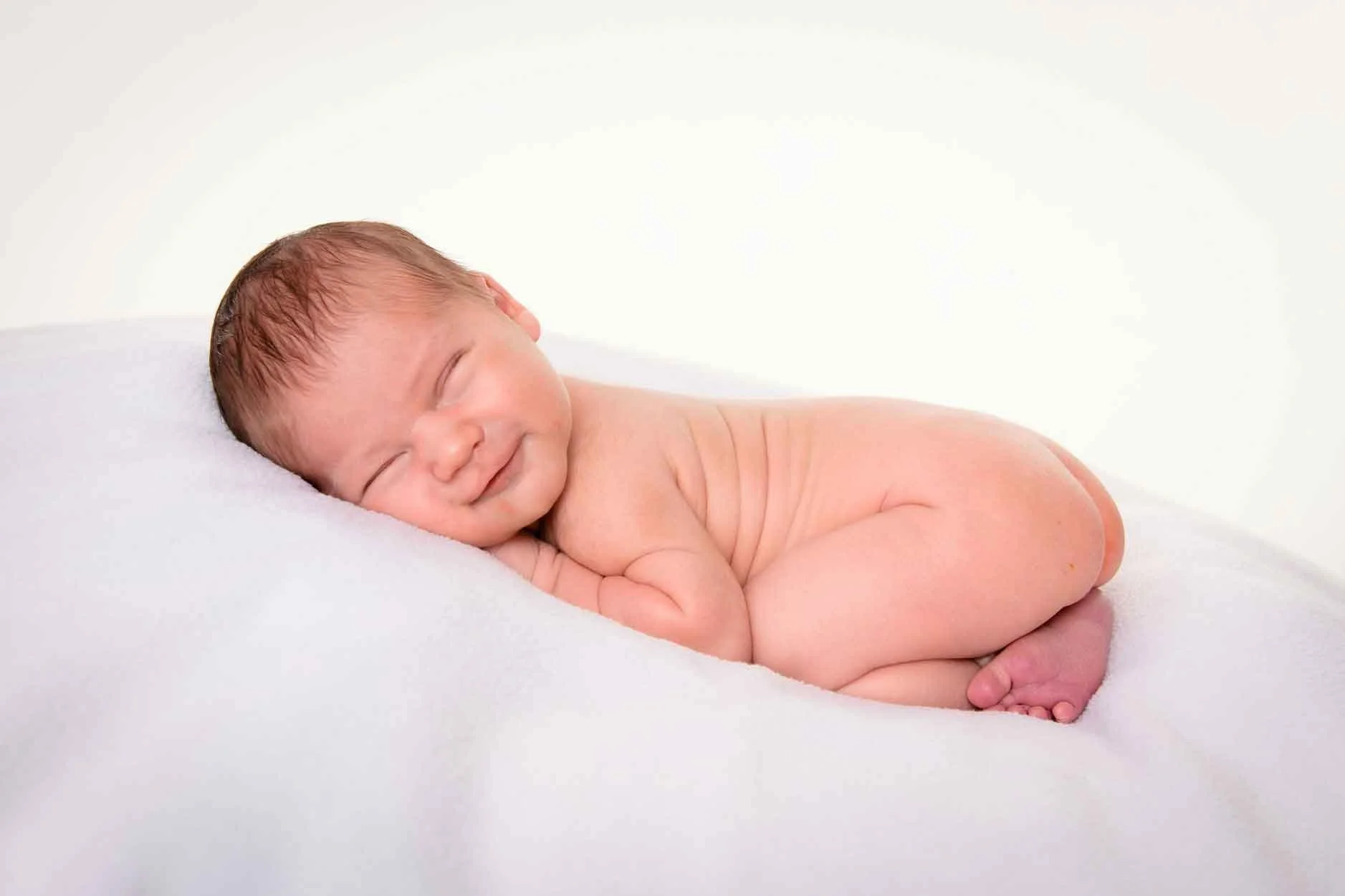 Nacktes Baby liegt auf einem weissen Kissen, lächelt entspannt und schläft.