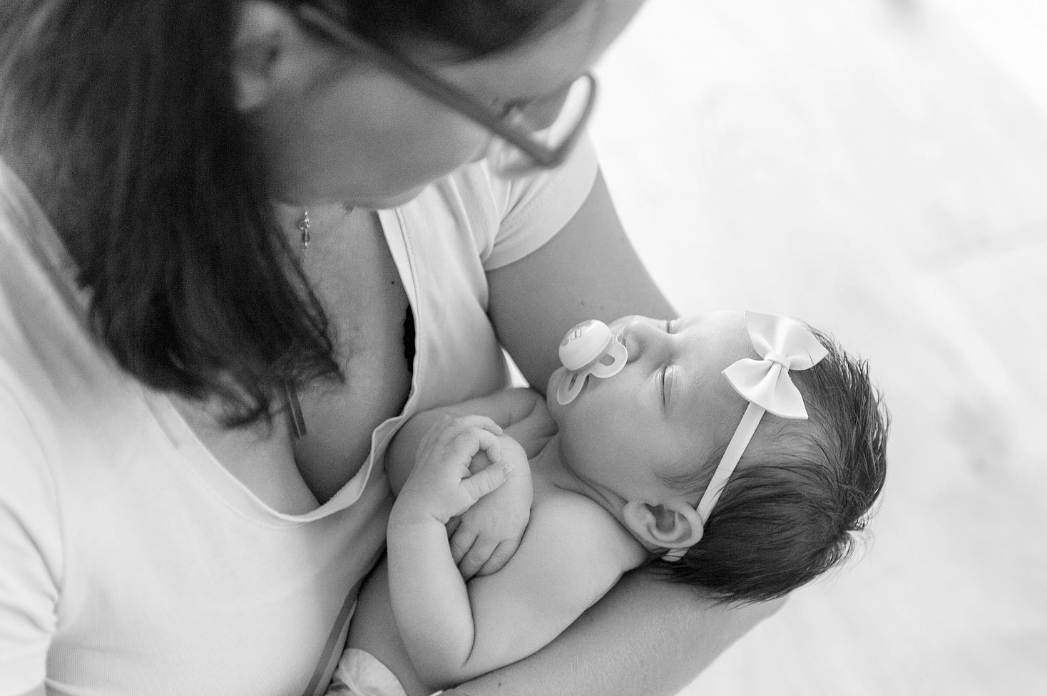 Schwarz-Weiss-Fotografie einer Person, die ein schlafendes Baby mit einer Schleife im Haar und einem Schnuller hält.