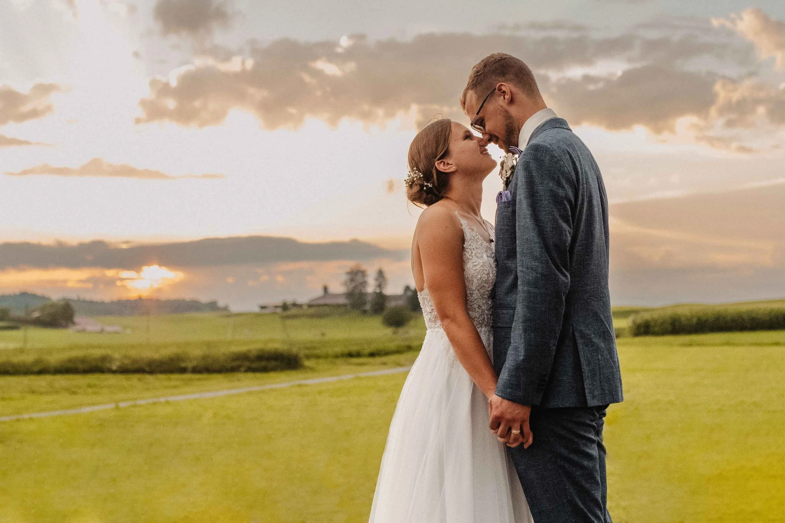 Brautpaar steht im Sonnenuntergang auf einer grünen Wiese und schaut sich liebevoll an, während ihre Stirn sich berühren.