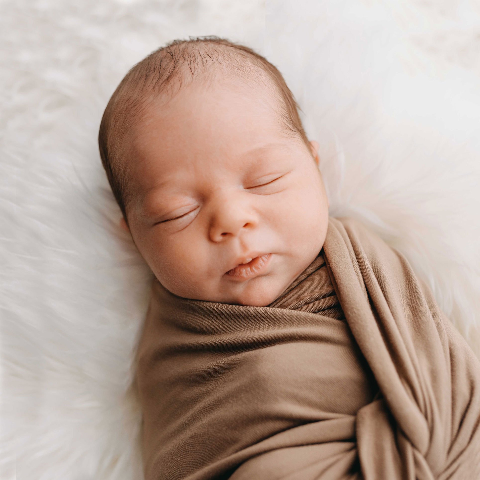 Schlafendes Baby eingewickelt in eine braune Decke auf einem weißen, flauschigen Untergrund.