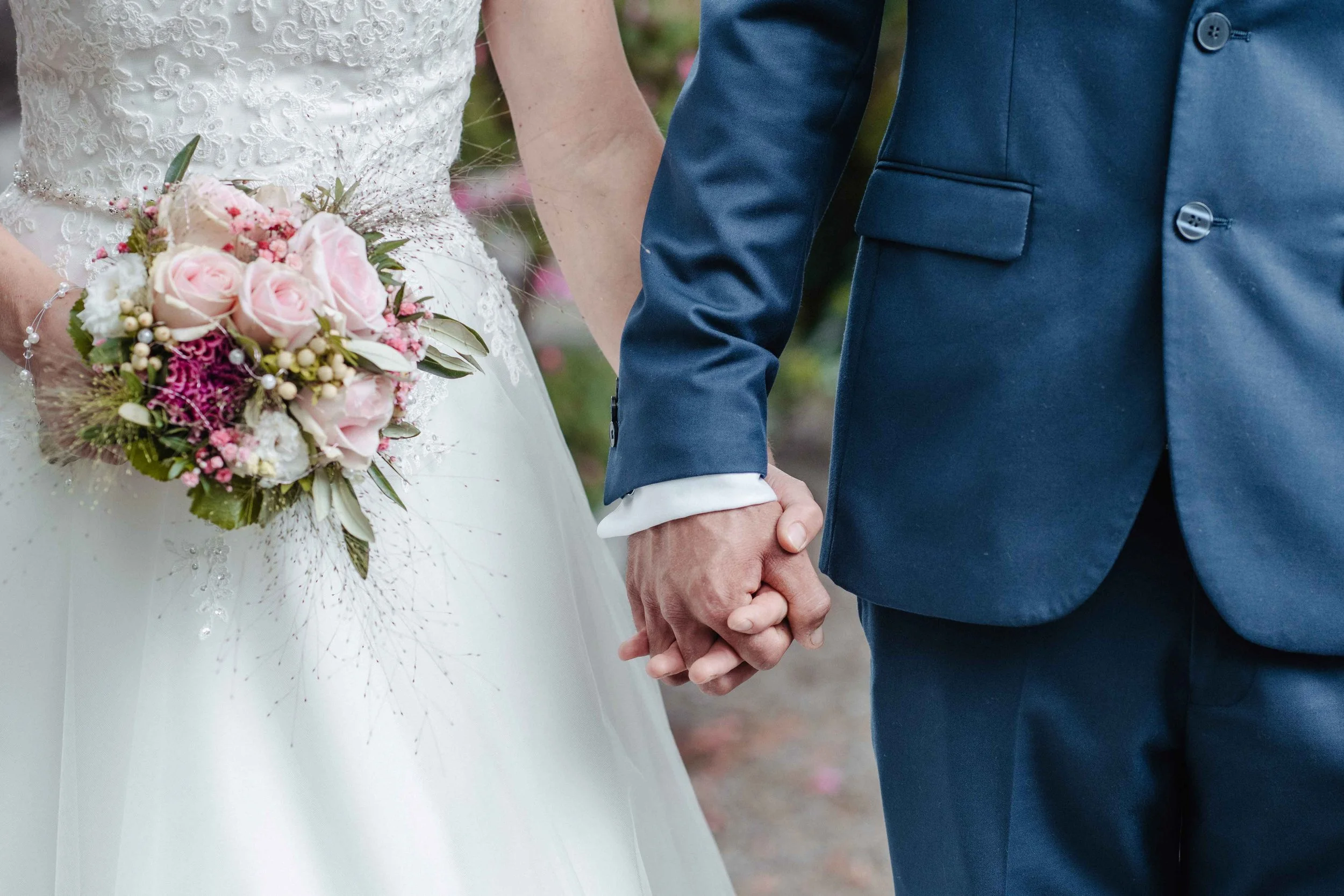 Brautpaar, das Händchen hält. Die Braut trägt ein weisses Hochzeitskleid und hält einen Brautstrauss mit rosa Rosen und anderen Blumen. Der Bräutigam trägt einen dunkelblauen Anzug.