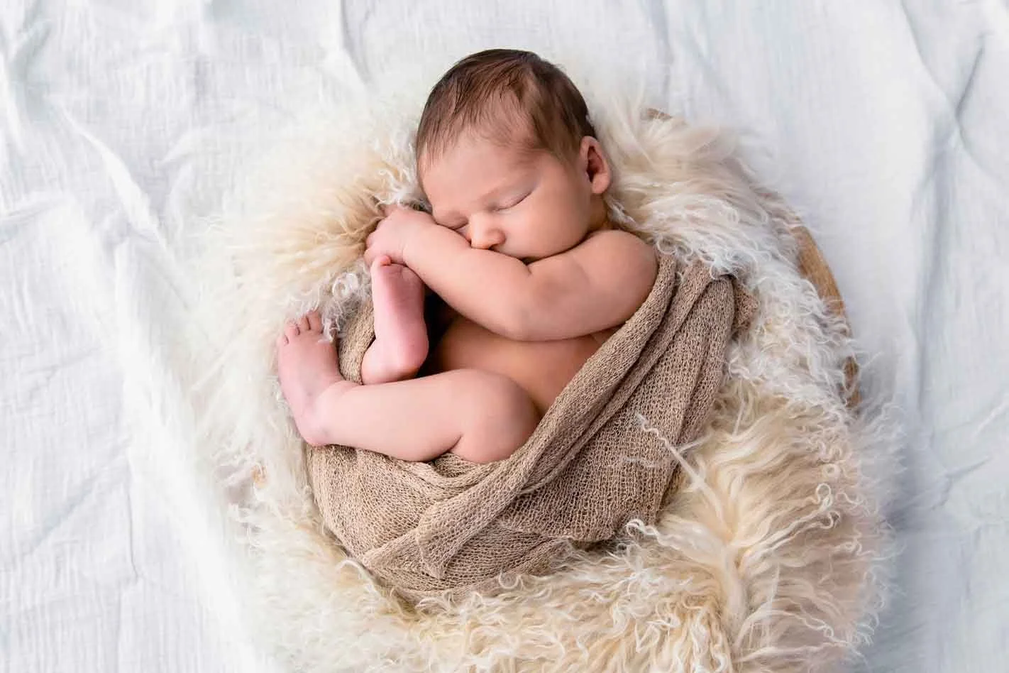 Ein schlafendes Neugeborenes, eingewickelt in einen braunen Stoff und auf einem flauschigen Fell liegend.