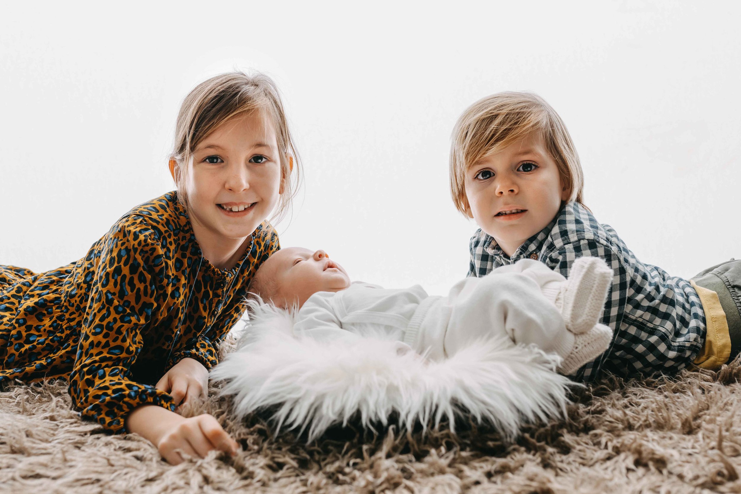 Drei Kinder liegen auf einem flauschigen Teppich, ein Baby in der Mitte, flankiert von einem Mädchen und einem Jungen, beide lächeln in die Kamera.