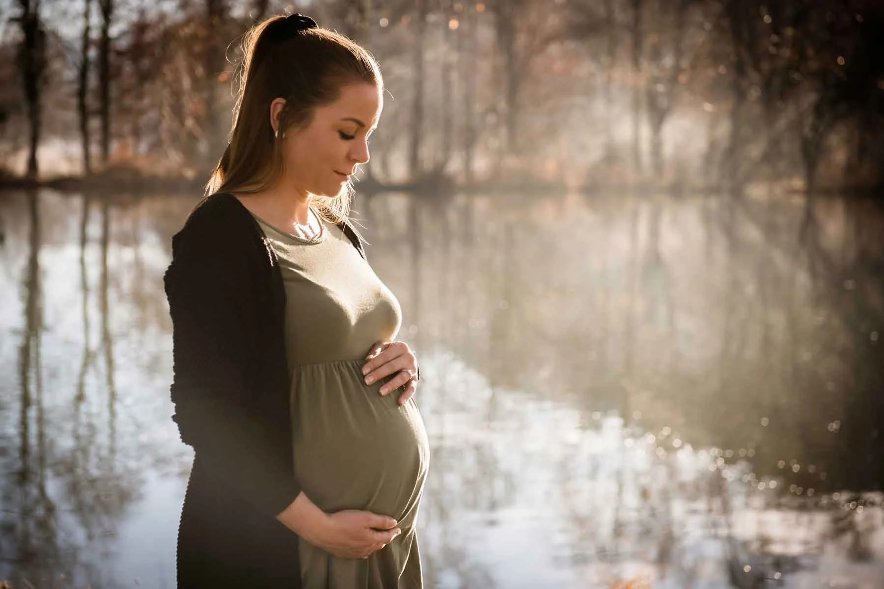 Schwangere Frau steht am See, hält ihren Bauch und sieht nach unten, umgeben von herbstlichen Bäumen.