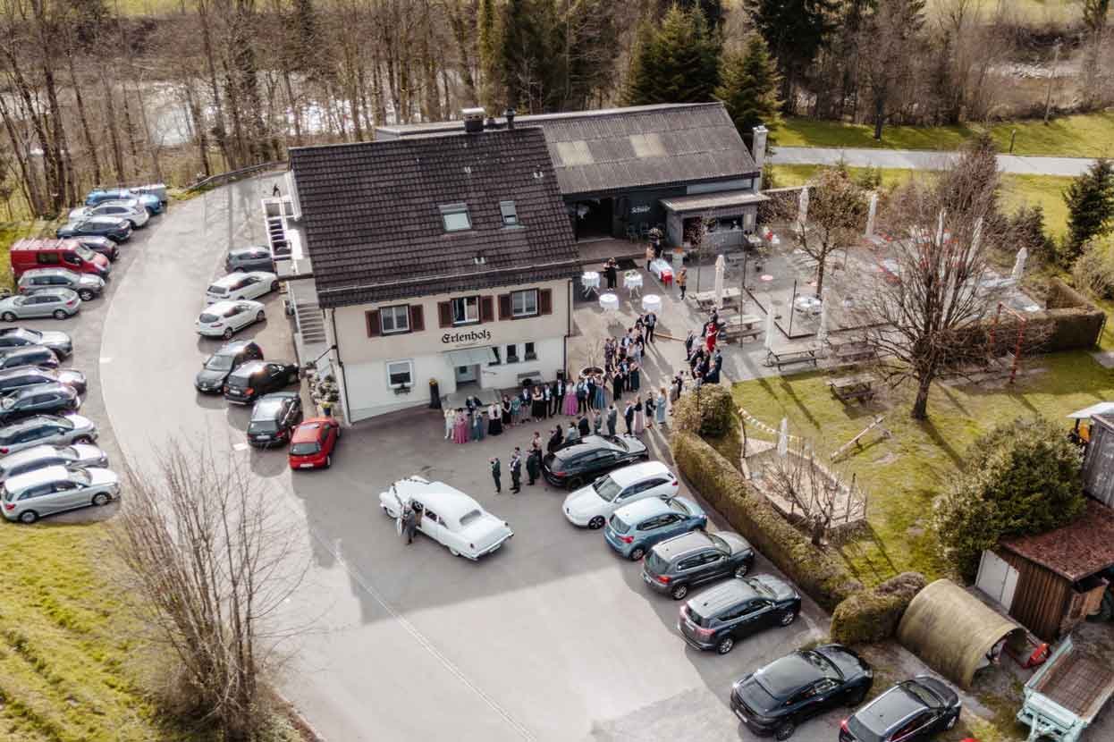 Gruppen von Menschen vor einem Restaurant mit Parkplätzen, Bäumen und Wäldern im Hintergrund warten auf das Brautpaar.