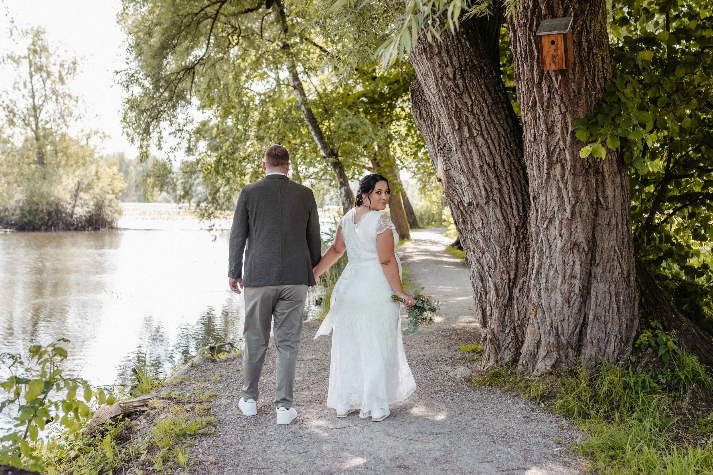 Brautpaar in Hochzeitskleidung hält Händchen an einem Weiher, umgeben von Bäumen, die Schatten spenden.