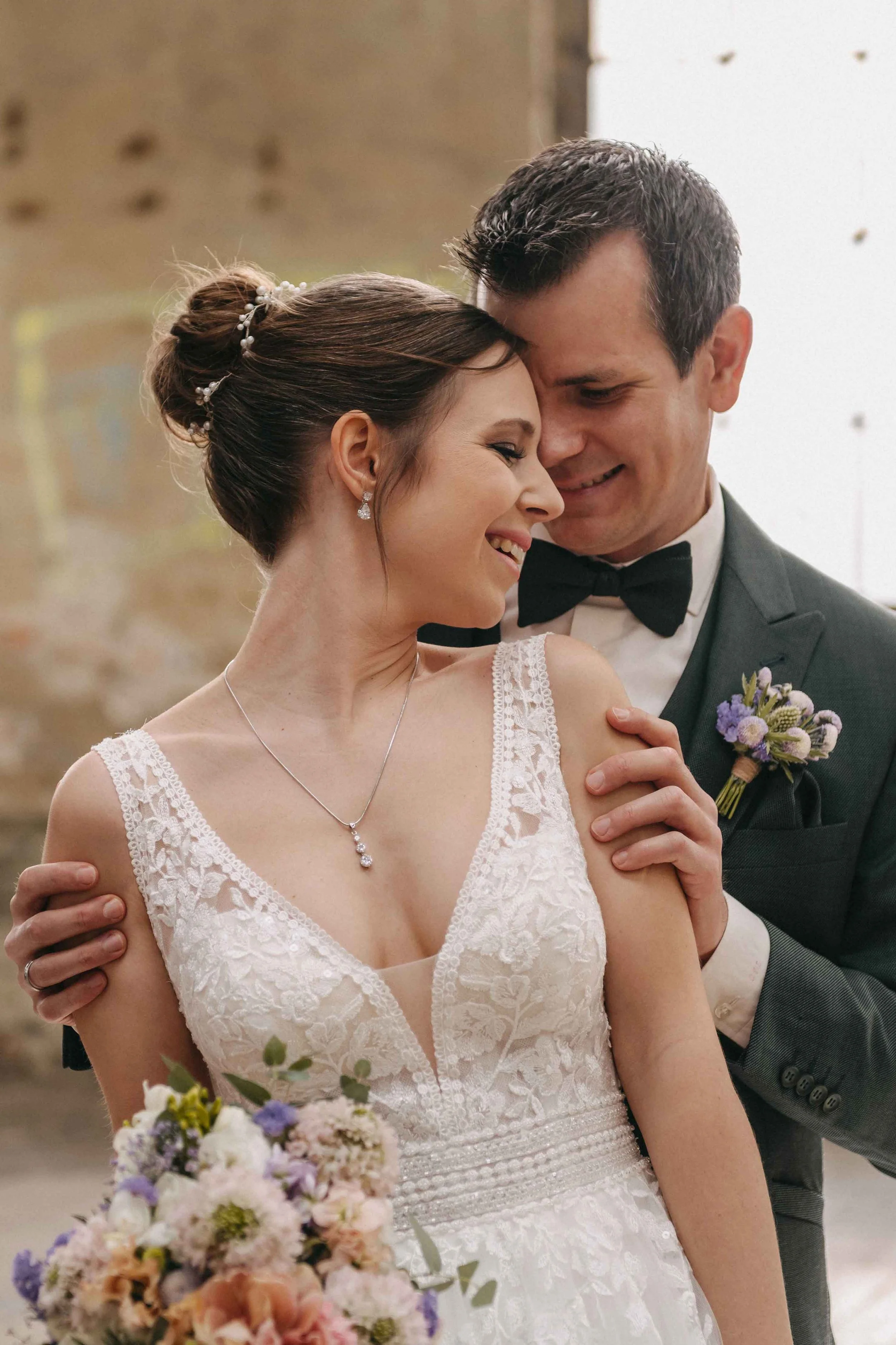 Glückliches Brautpaar, die Frau trägt ein weisses Spitzenkleid mit Schmuck, der Mann trägt einen dunklen Anzug mit Fliege, beide lächeln sich an.