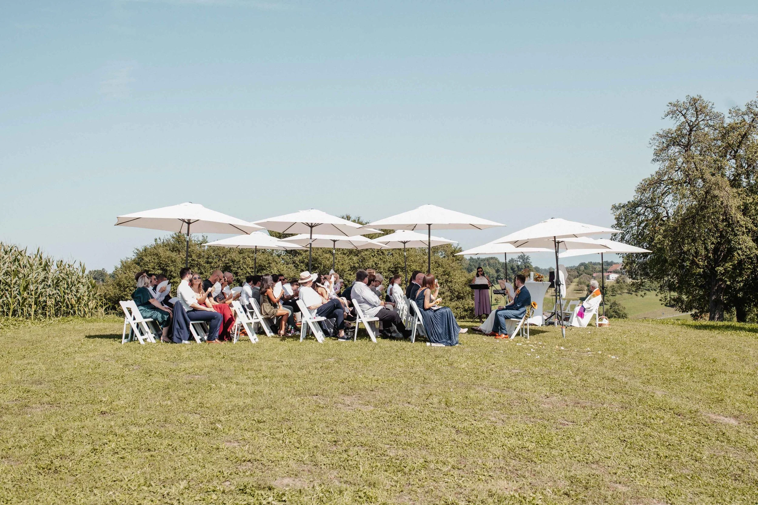 Eine Gruppe von Menschen bei einer Outdoor-Hochzeit im Grünen unter weissen Sonnenschirmen, mit einem Rednerpult und Musikern im Hintergrund.