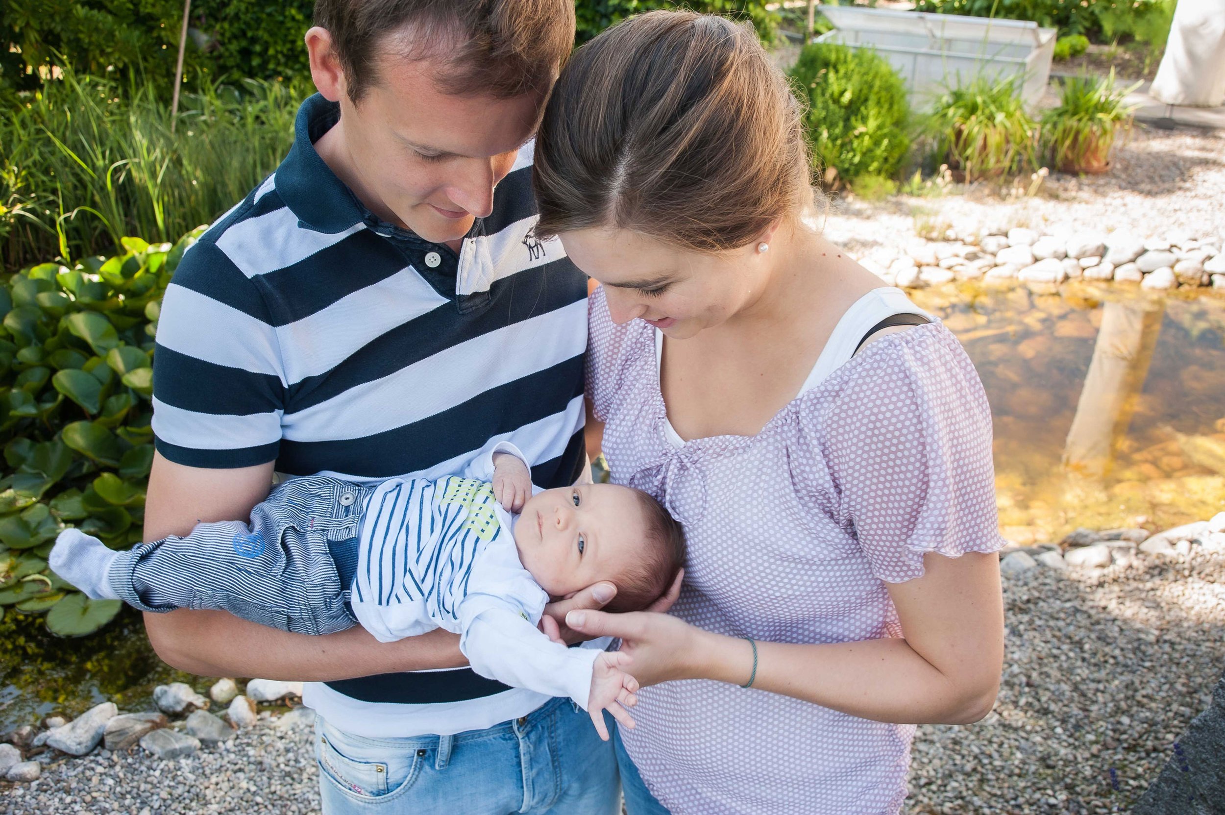 Ein Paar hält ein Baby im Freien. Der Mann trägt ein gestreiftes Polohemd, die Frau ein gepunktetes Oberteil. Das Baby liegt in ihren Armen. Im Hintergrund ist ein Teich und Grünpflanzen zu sehen.