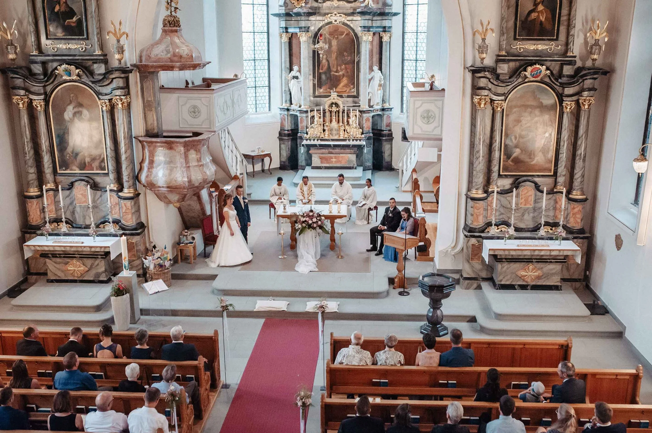 Hochzeitszeremonie in einer Kirche mit Braut, Bräutigam, Pfarrer und Gästen, mit traditioneller Inneneinrichtung und Altar im Hintergrund.