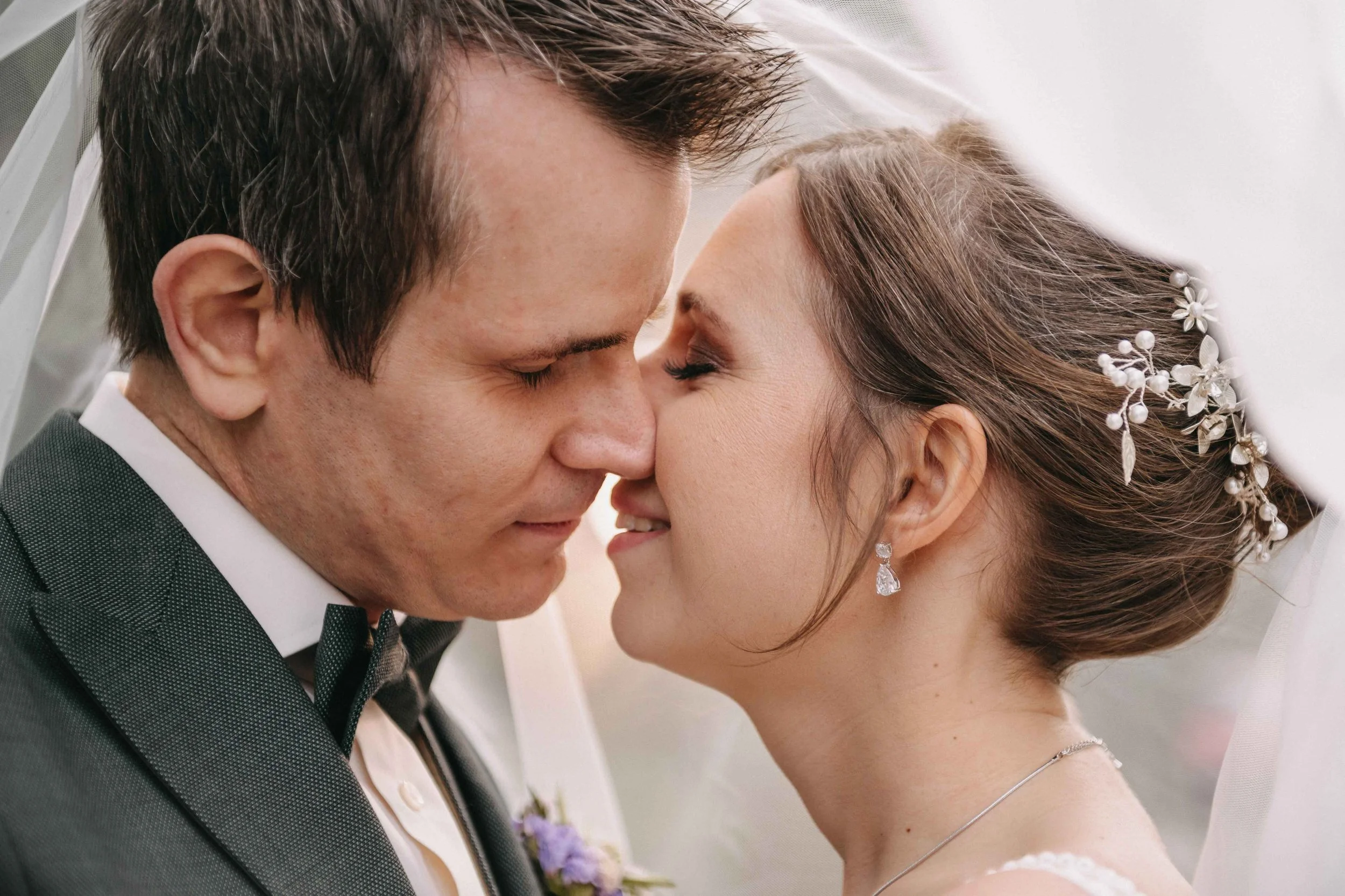 Brautpaar bei einer Hochzeit, völlig nah beieinander mit ihrer Stirn und Nase berührend, lächelnd, mit vorbereiteten Haaren und Schmuck, unter einem weissen Schleier.