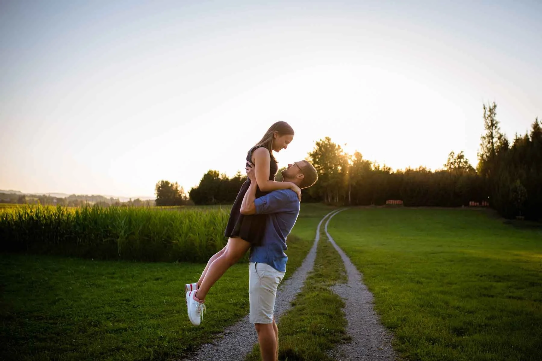 Ein Paar auf einer ländlichen Strecke bei Sonnenuntergang, der Mann hebt die Frau hoch, die lachen und sich umarmt.