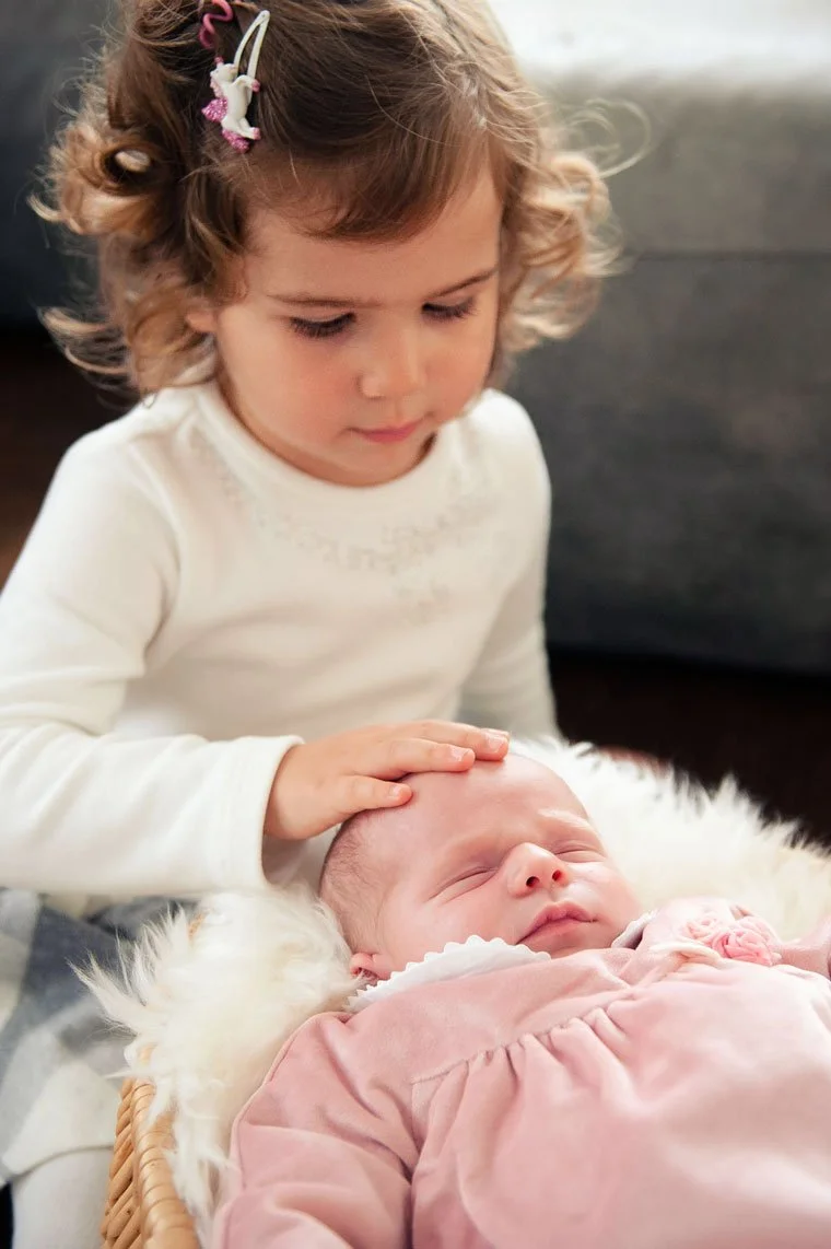 Ein kleines Mädchen das eine schlafende Baby auf einem weichen, pelzigen Untergrund berührt.