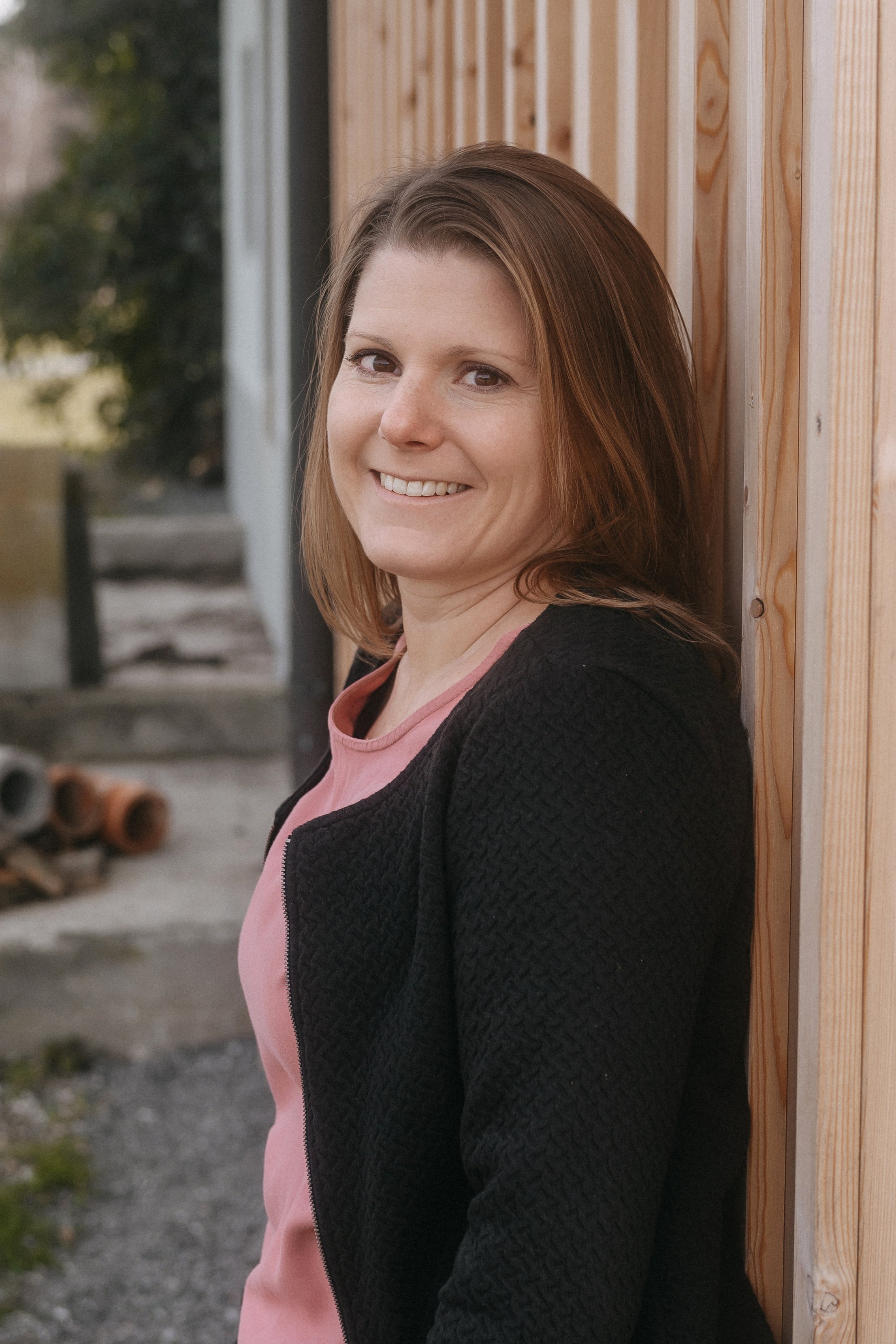 Janine Huber lehnt an Holzwand, lächelt, trägt rosa Shirt und schwarze Jacke, Hintergrund unscharf.