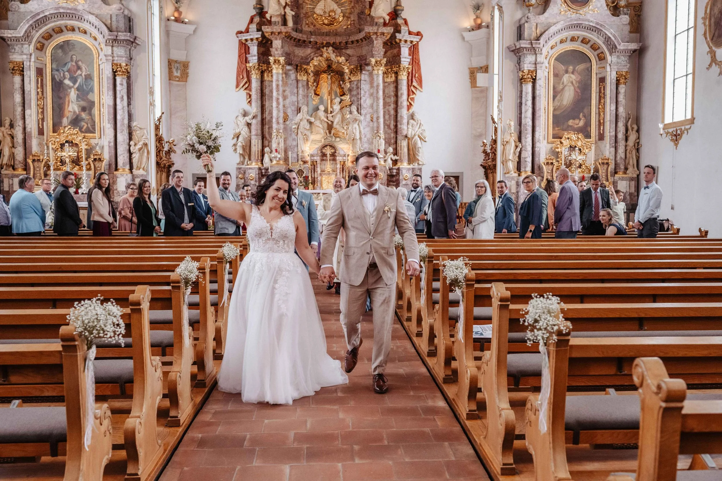 frisch verheiratetes Brautpaar, gehen Hand in Hand durch die Kirche, umrahmt von Gästen. Die Kirche ist reich verziert mit goldenen und barocken Elementen, großem Altar.