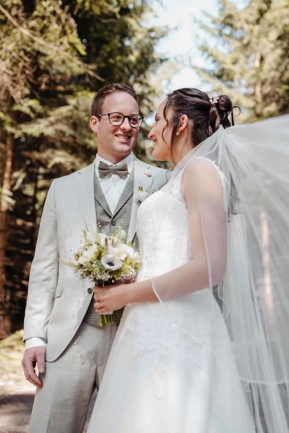 Hochzeitspaar steht in einem Wald, die Braut hält den Brautstrauss und trägt ein weisses Hochzeitskleid, der Bräutigam trägt einen grauen Anzug mit Weste. Sie schauen sich liebevoll an.