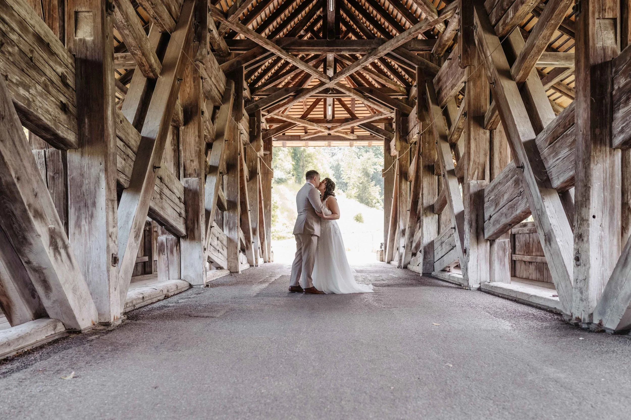 Brautpaar in einer überdachten Holzbrücke, romantische Umgebung mit Sonnenlicht im Hintergrund.